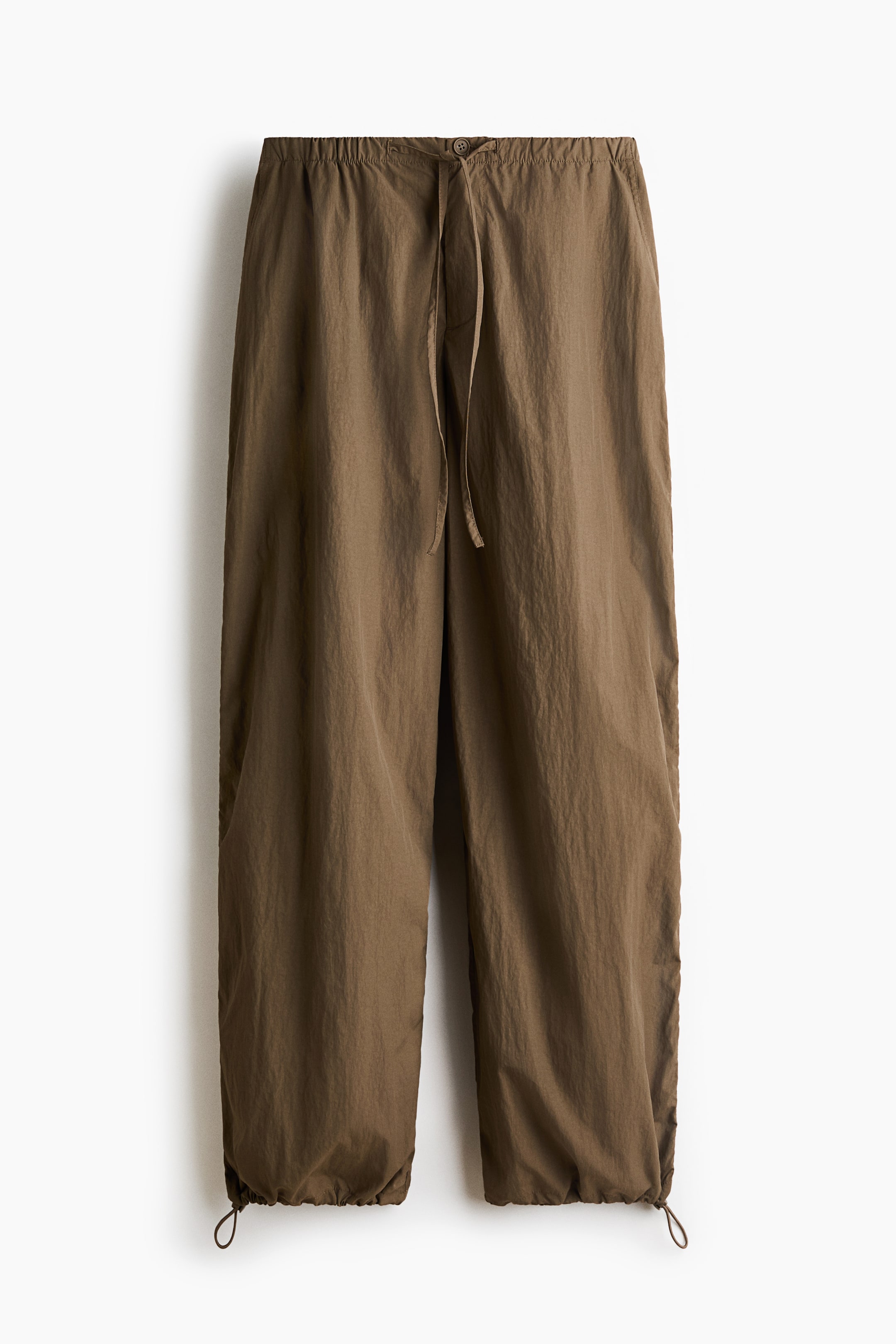 View larger image: A pair of brown nylon trousers is hanging front-facing, featuring a relaxed fit silhouette with an elasticated drawstring waistband and button closure. The slightly tapered legs extend down to adjustable hems with visible drawstring loops.
