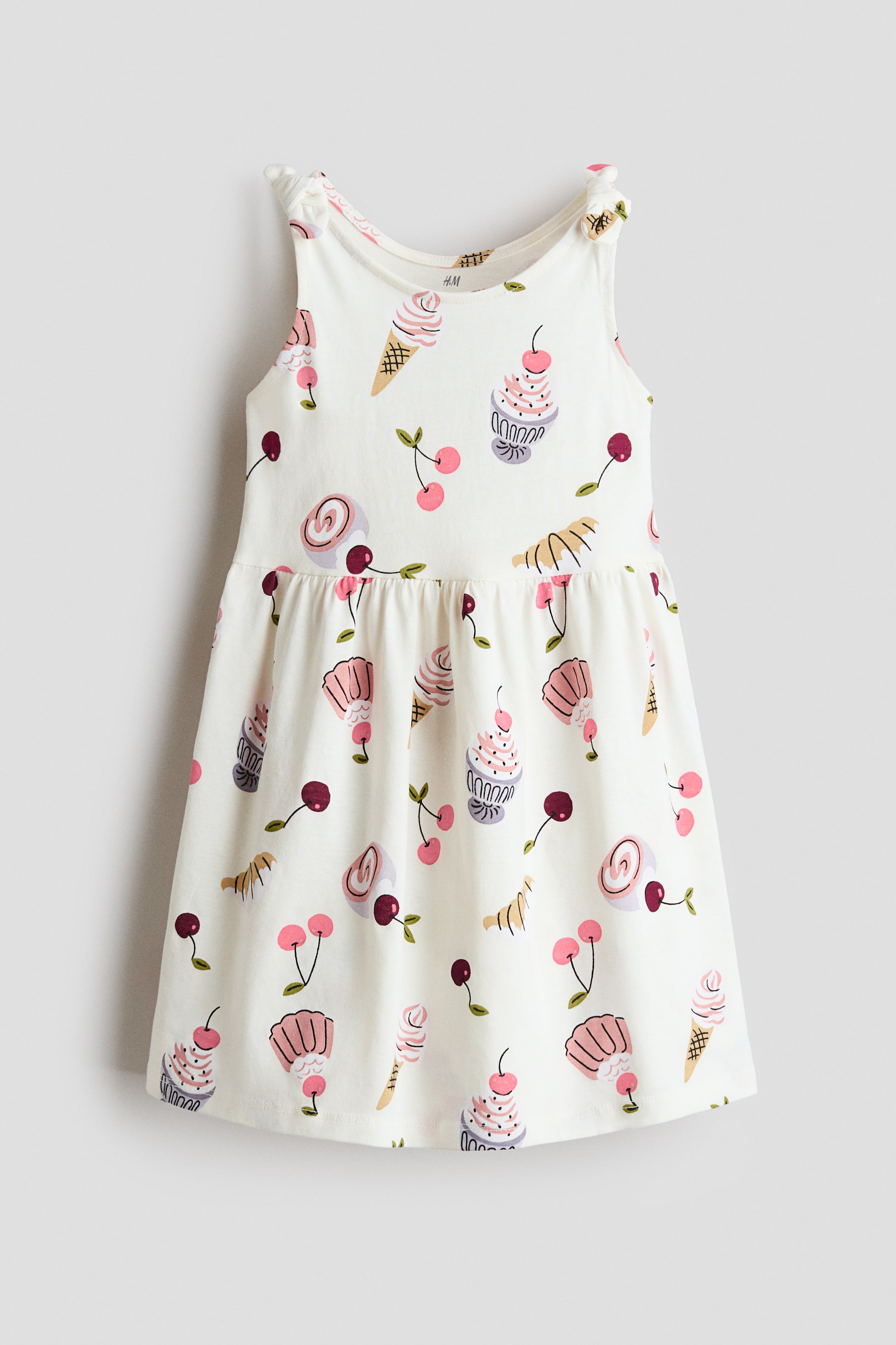 View larger image: The sleeveless dress is showcased as a cream-coloured garment, facing front. It features a round neckline and knotted details on the shoulder straps, while an all-over print of ice cream cones, cupcakes, pastries, and cherries embellishes the fabric. A gathered waistline leads to a flaring skirt.