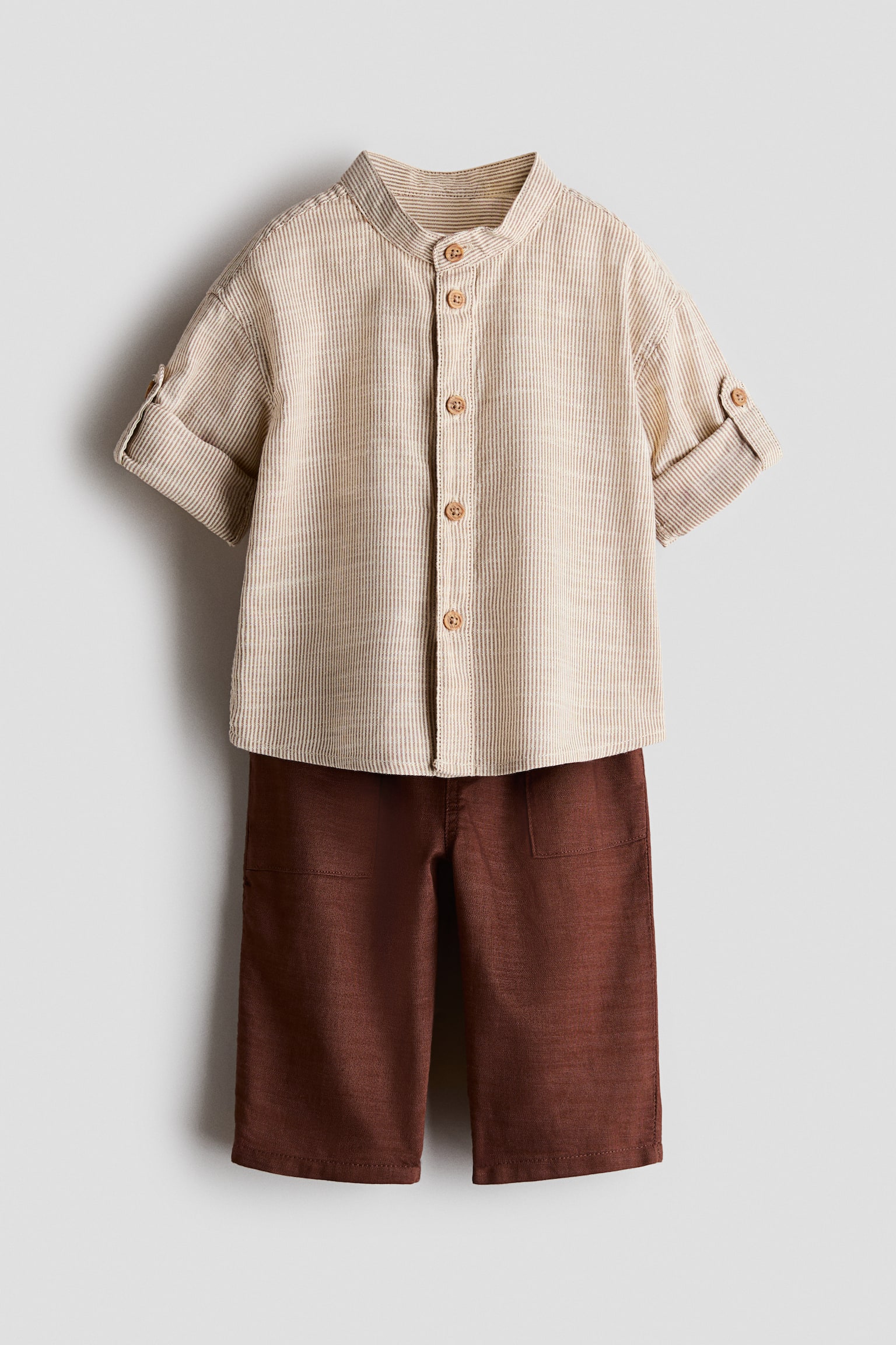 A two-piece clothing set is shown front-facing, featuring a beige and white vertically striped shirt and dark brown trousers. The shirt features a band collar, a front button closure, and short sleeves with rolled cuffs secured by button tabs, while the trousers feature a straight-leg fit with front patch pockets.