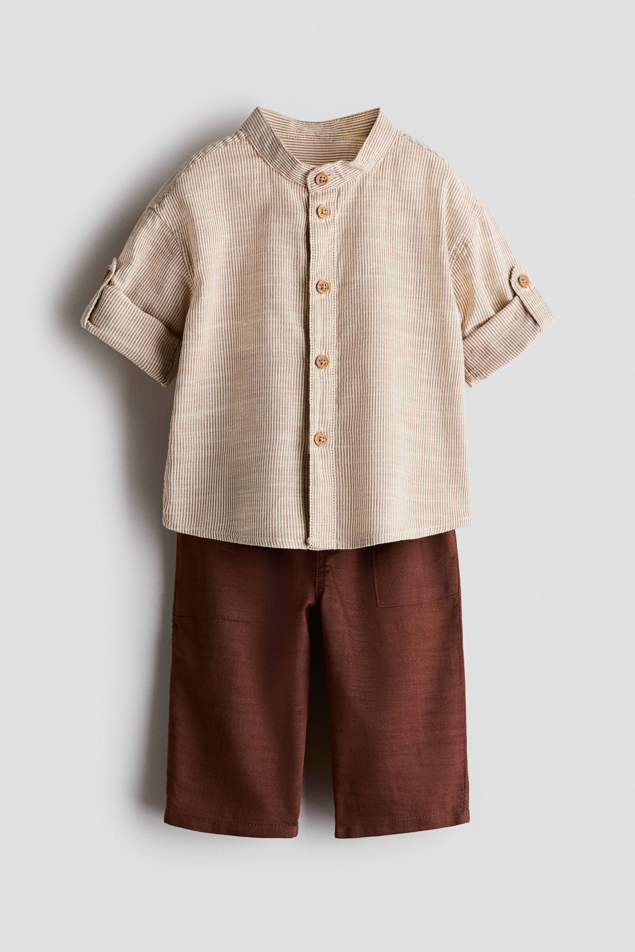 View larger image: A two-piece clothing set is shown facing front, featuring a beige and white vertically striped shirt and dark brown trousers. The shirt displays a band collar, a front button closure, and short sleeves with rolled cuffs secured by button tabs, while the trousers feature a straight-leg fit with front patch pockets.