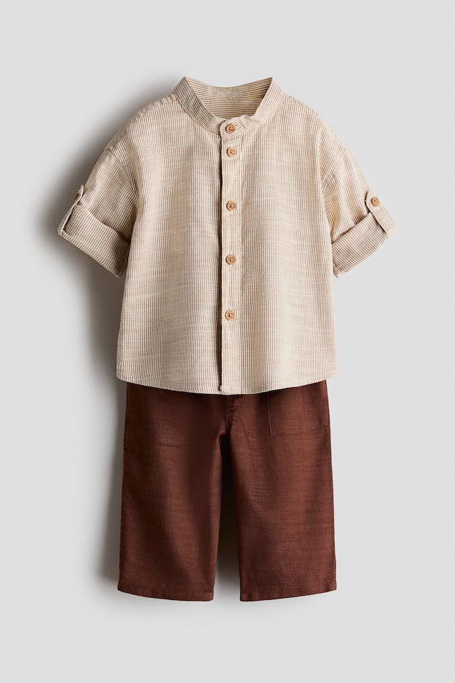 A two-piece clothing set is shown from the front, featuring a beige and white vertically striped shirt and dark brown trousers. The shirt features a band collar, a front button closure, and short sleeves with rolled-up cuffs secured by button tabs, while the trousers feature a straight-leg fit with front patch pockets. - 1