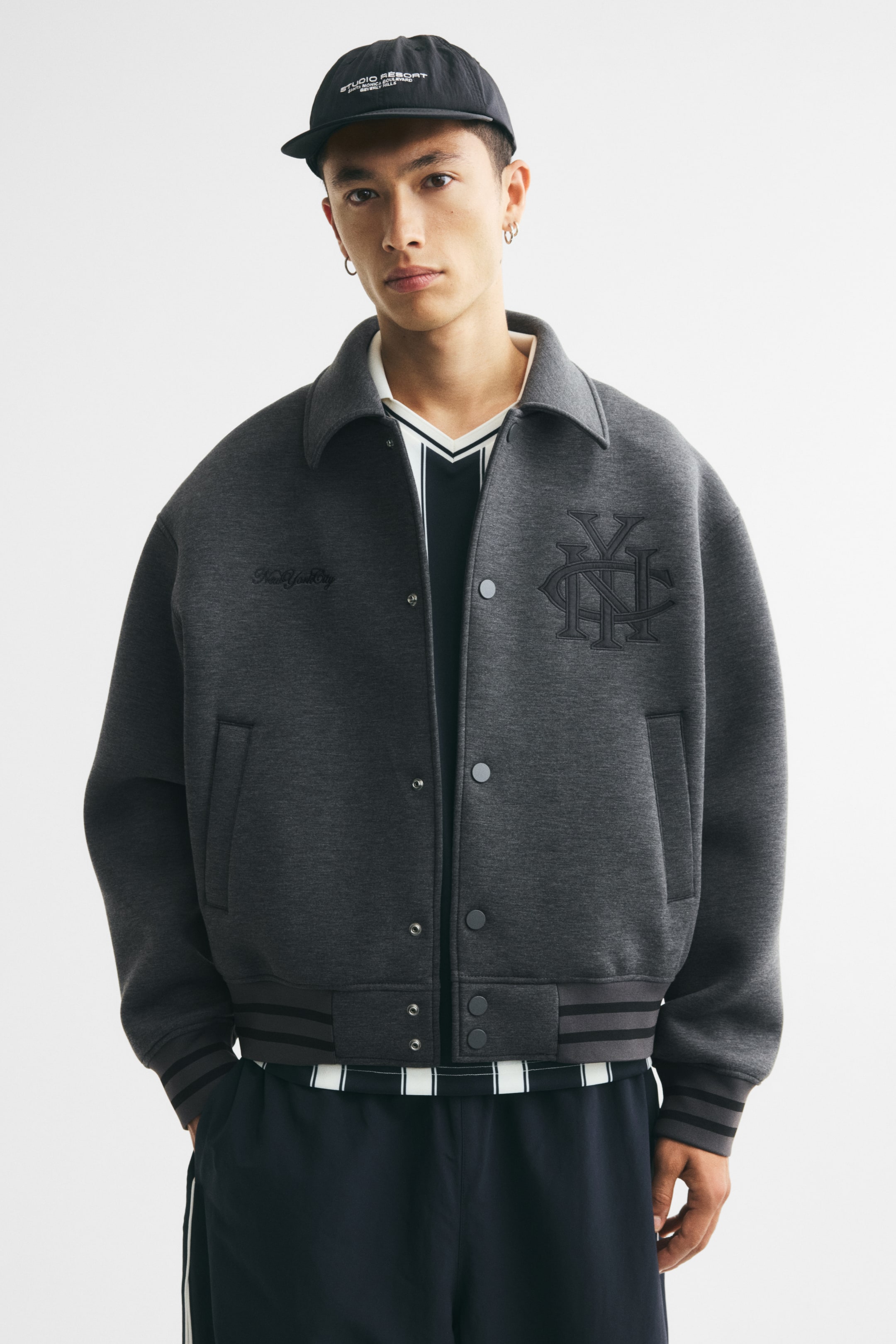 Agrandir l'image: Une veste de baseball gris foncé en tissu scuba est portée ouverte par le mannequin, mettant en valeur son col, sa fermeture à boutons-pression et ses motifs brodés sur la poitrine. La veste présente des poches passepoilées et des finitions côtelées rayées, portée sur une chemise rayée à col V et un pantalon foncé.