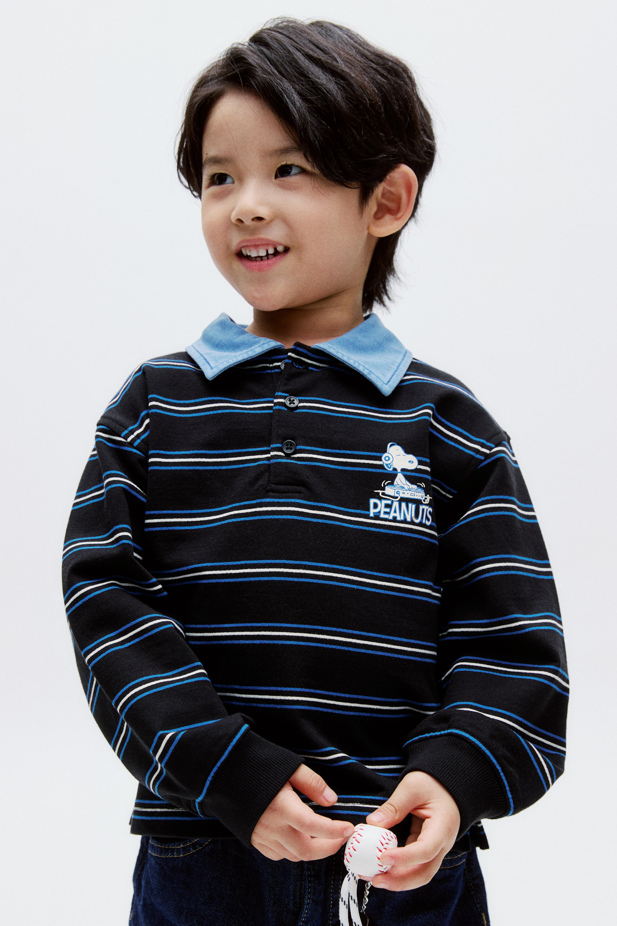 拡大画像を表示: 黒の長袖ポロシャツは、青と白の細い横縞が特徴で、コントラストの効いた青い襟とリブ付きカフスが付いています。首元には2つボタンのプラケットが見え、左胸には「PEANUTS」の文字とスヌーピーのアビエーターグラフィックが飾られています。