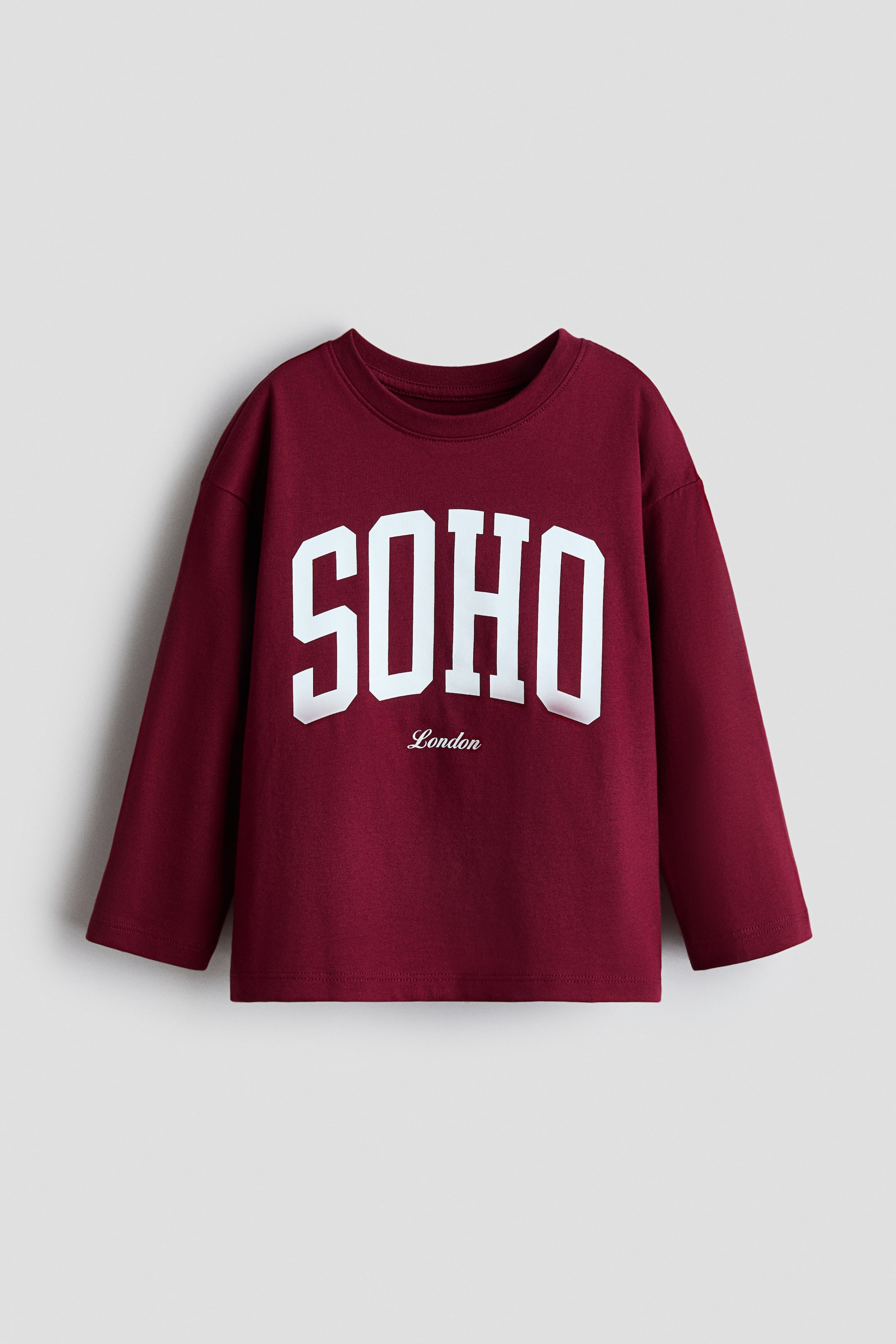 View larger image: A maroon long-sleeved top is showcased front-facing, featuring a crew neck and a straight hem. Large white block letters spelling 'SOHO' are displayed on the chest, with 'London' in smaller white script text positioned beneath.