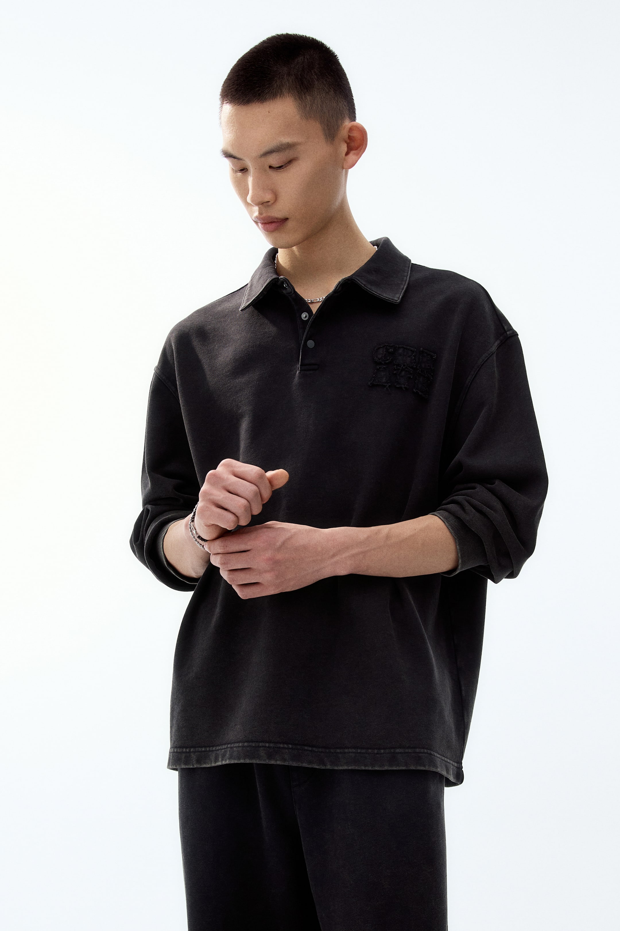 이미지 확대 보기: 워시드 블랙 컬러의 긴소매 폴로셔츠로, 약간 비스듬한 정면 각도에서 촬영되어 클래식 칼라와 투 버튼 플래킷을 갖추고 있습니다. 오버사이즈 핏에 골지 소매단이 적용되었으며 왼쪽 가슴에는 어둡고 텍스처감 있는 아플리케 장식이 있습니다.