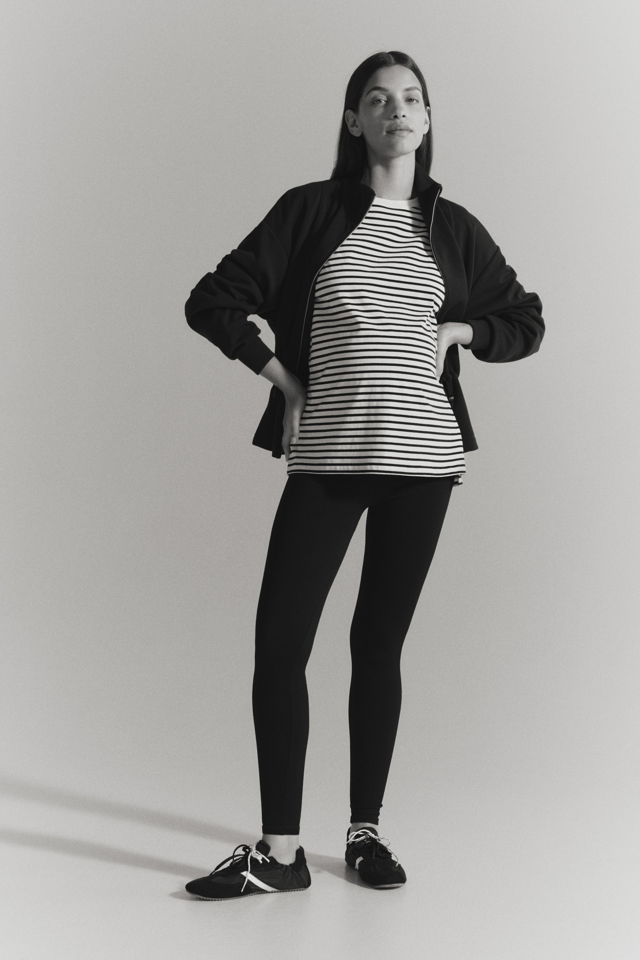 Ver imagem maior: A modelo está a usar leggings pretas em jersey canelado, com as mãos na cintura. As leggings até ao tornozelo estão combinadas com uma t-shirt às riscas, um casaco preto aberto e ténis.