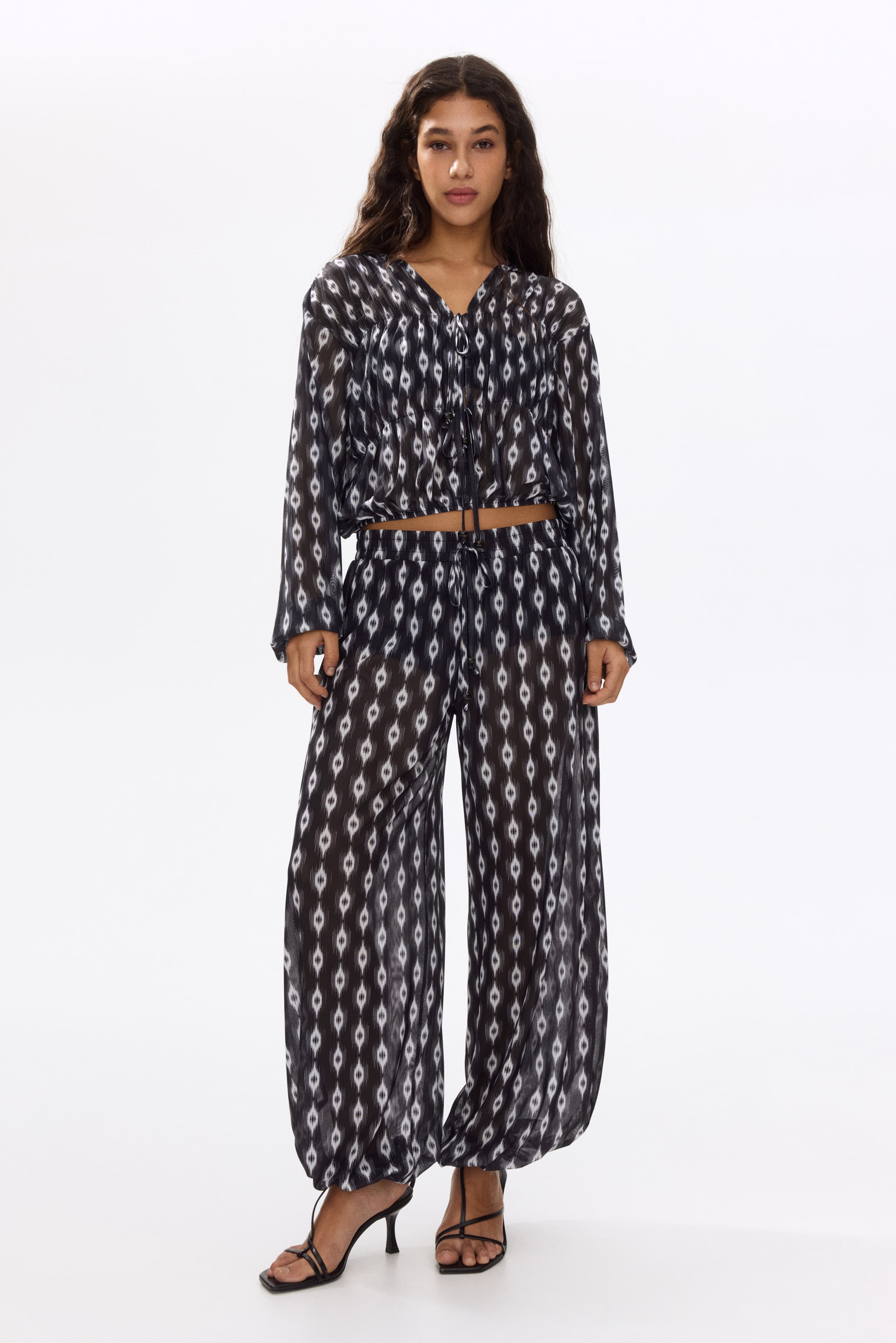 View larger image: The black and white sheer set features a top and matching wide-leg pants, both showcasing an ikat-style diamond pattern. The top is crafted with a V-neckline, a front drawstring tie, long sleeves, and a drawstring hem, while the pants boast an elasticated drawstring waistband and gathered elasticated hems.
