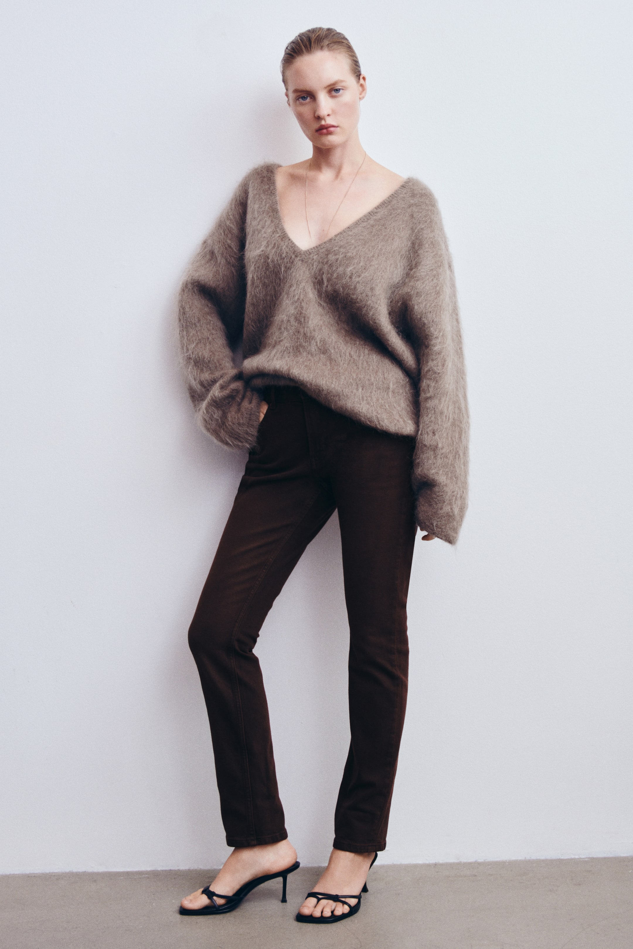 Agrandir l'image: Le modèle porte un jean slim-fit marron foncé, en se tenant debout avec une main posée dans la poche avant. Le jean présente une coupe skinny le long des jambes et est associé à un pull taupe oversize à col V, duveteux, ainsi qu’à des sandales noires à talons.