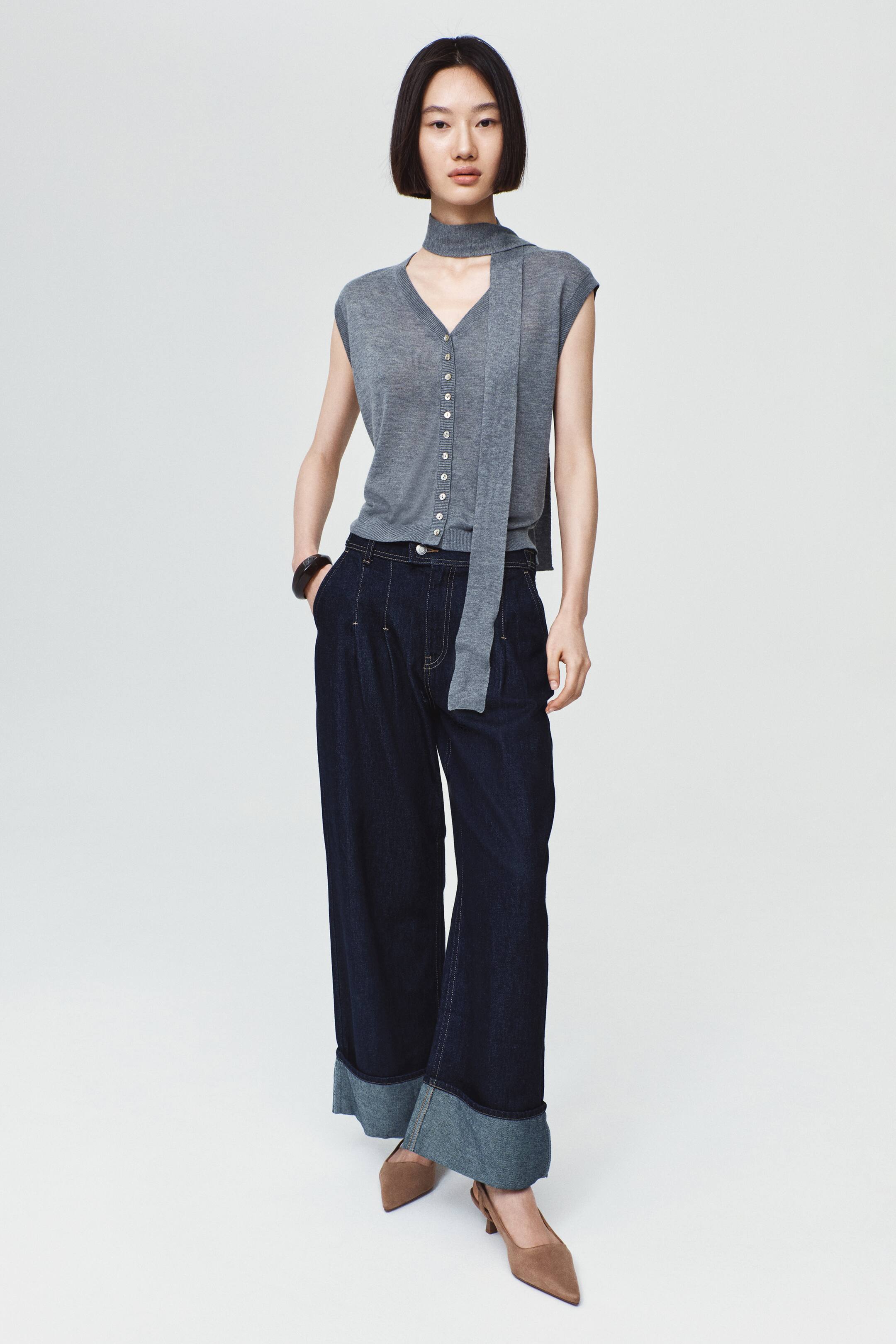 View larger image: Dark-wash denim jeans with a wide-leg silhouette, front pleats, and wide turned-up cuffs are worn by the model. The jeans are styled with a sleeveless grey knit top and brown pointed-toe heels, with the model standing and resting one hand in a pocket.