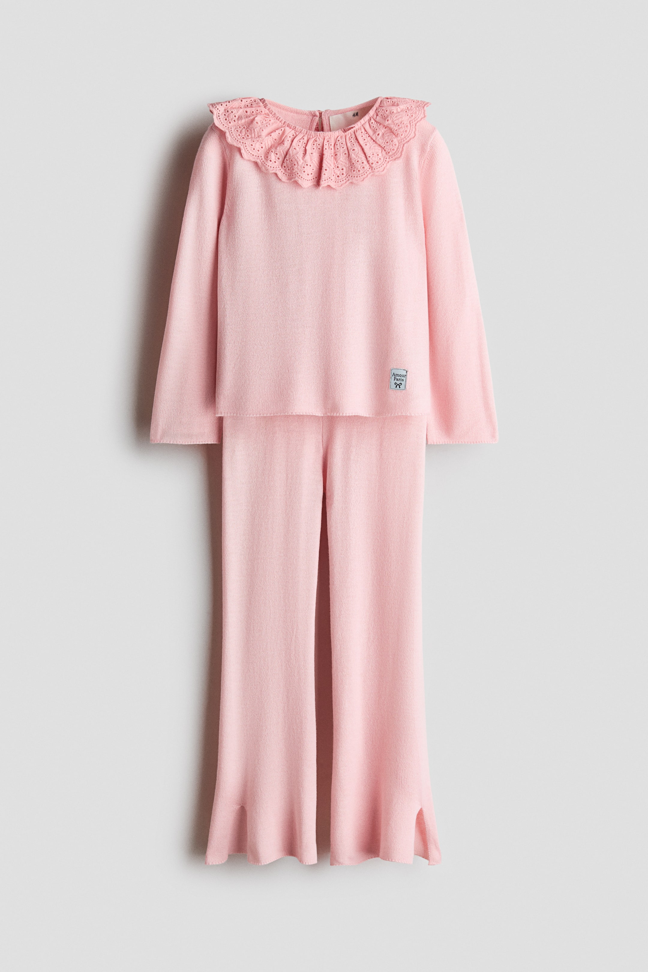Größeres Bild anzeigen: Ein weiches, feines Strick-Set in Rosa präsentiert ein langärmeliges Oberteil und eine passende Hose, von vorne gezeigt. Das Oberteil verfügt über einen runden, elastischen Halsausschnitt mit einer breiten, gewellten Broderie-Anglaise-Stufe sowie einen Tropfenausschnitt mit Knopf am Rücken. Die Hose zeigt einen elastischen Bund mit ausgestellten Beinen und Schlitzen am Saum.