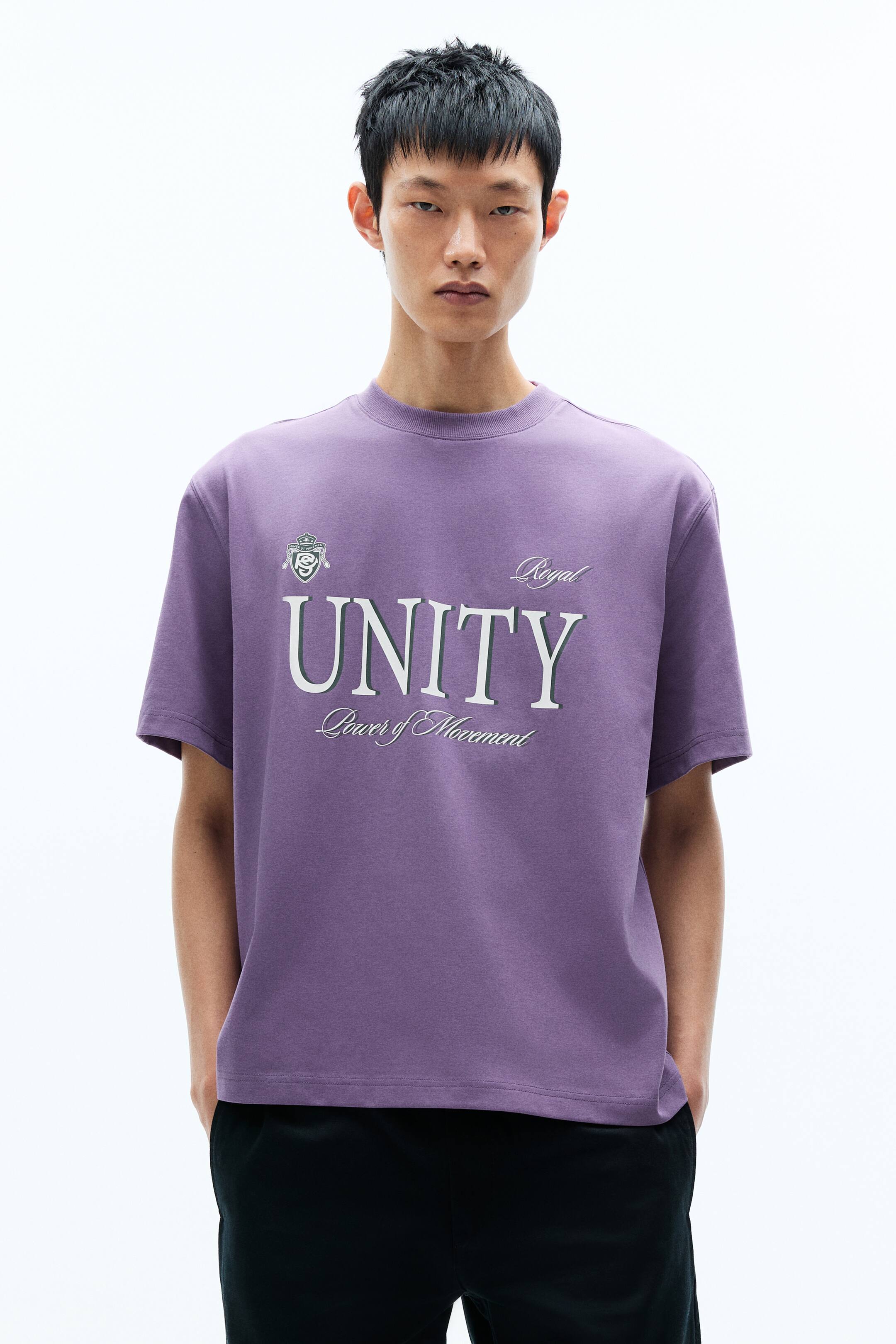 View larger image: Purple crew-neck T-shirt with short sleeves and a relaxed fit. The front features a large white graphic with dark outlining that reads "UNITY", accompanied by "Royal" in script above and "Power of Movement" in script below, plus a crest on the upper-left chest.