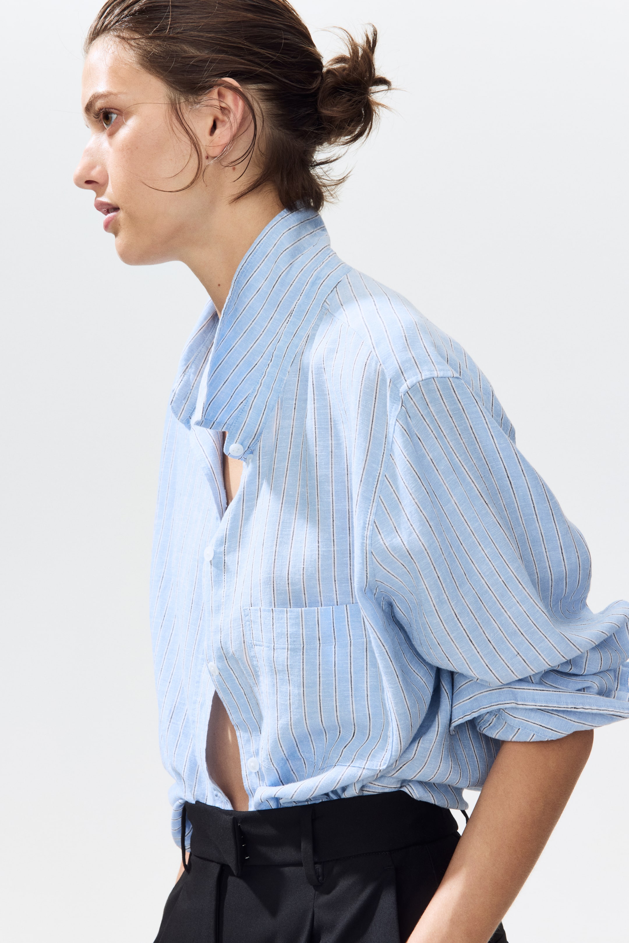 查看大圖: 淺藍色的扣鈕襯衫配有細密的深色垂直條紋，從側面角度展示。這款襯衫採用寬鬆的休閒版型，左胸設有口袋，全袖在袖口處聚攏，部分鈕扣未扣，搭配黑色長褲。