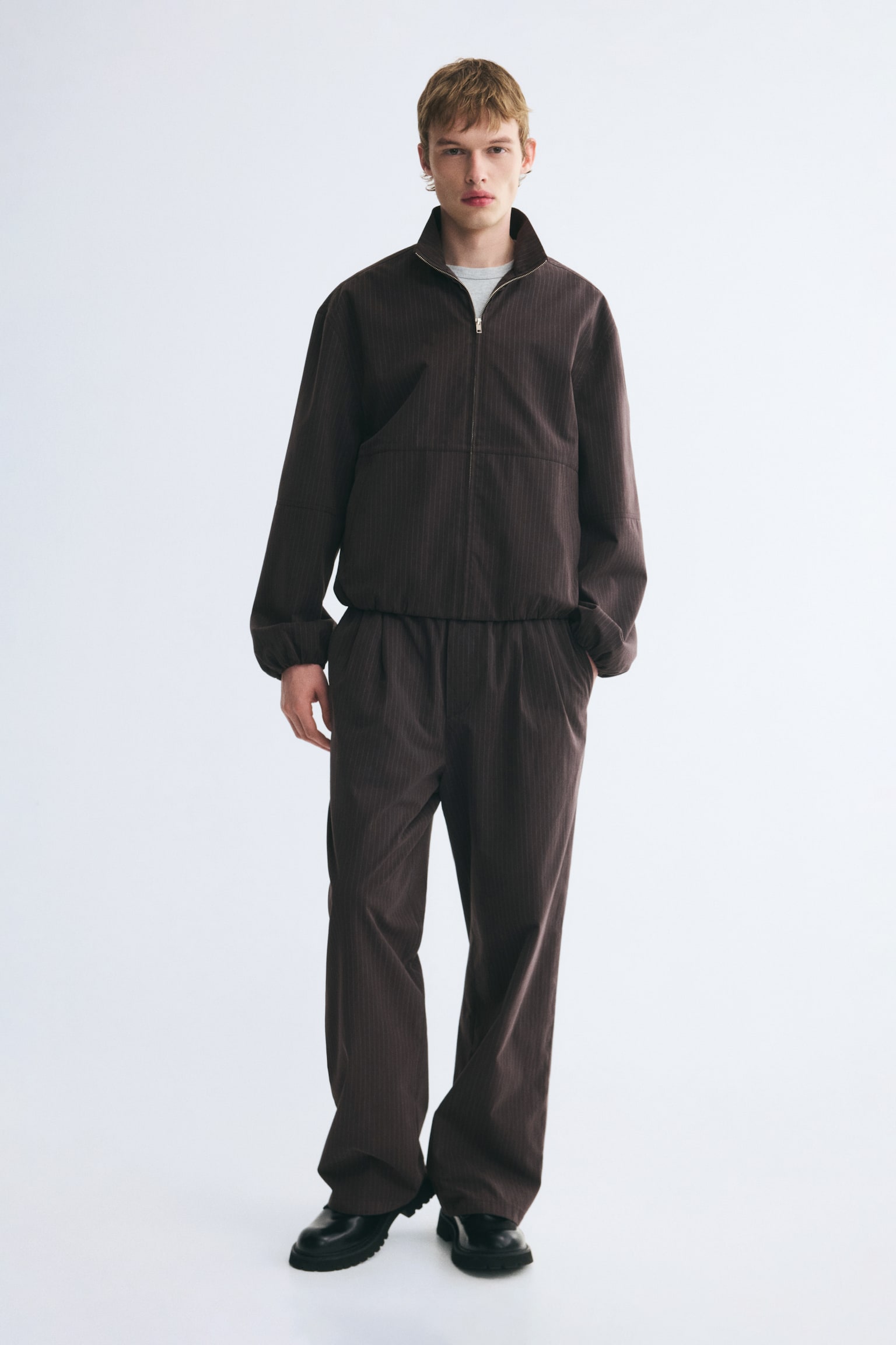 Dunkelbraune Hosen aus Baumwollmischung mit lockerer Passform, Bundfalten vorne und geradem Bein werden vom Model getragen. Die Hose, kombiniert mit einer passenden Jacke mit ReiÃverschluss und schwarzen Schuhen, zeigt eine entspannte Silhouette, wÃ¤hrend das Model eine Hand lÃ¤ssig in einer schrÃ¤gen Seitentasche ablegt. - 1