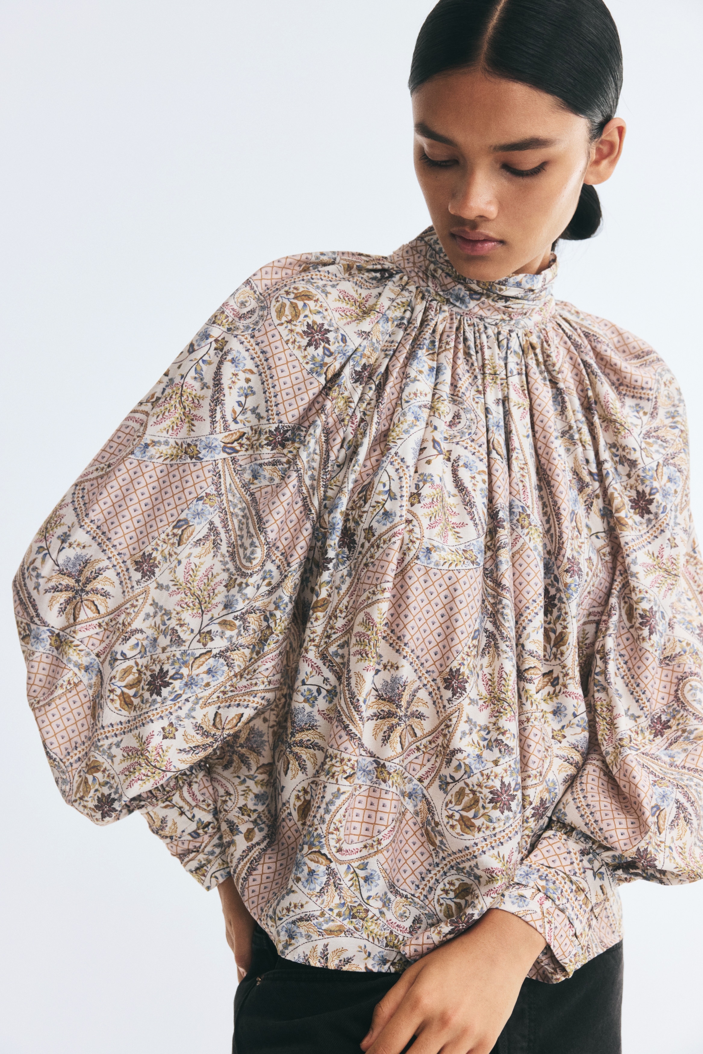 Esta vista frontal muestra una blusa de corte oversize en un tejido de sarga con un intrincado estampado paisley multicolor. La blusa presenta un cuello alto de tirilla fruncida con frunces en la base, que cae sobre un cuerpo voluminoso con largas mangas raglán abullonadas y puños anchos.