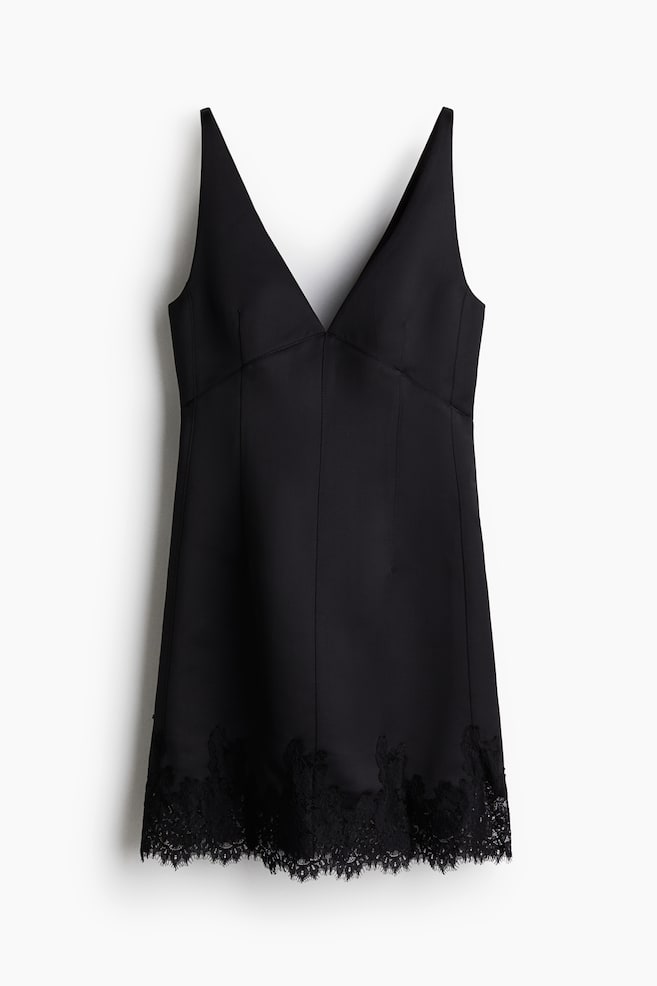 Une robe noire mini est présentée de face, avec un décolleté en V profond et un corsage structuré avec une couture horizontale sous la poitrine et des coutures verticales. Son ourlet droit est fini avec une bordure en dentelle noire complexe. - 1