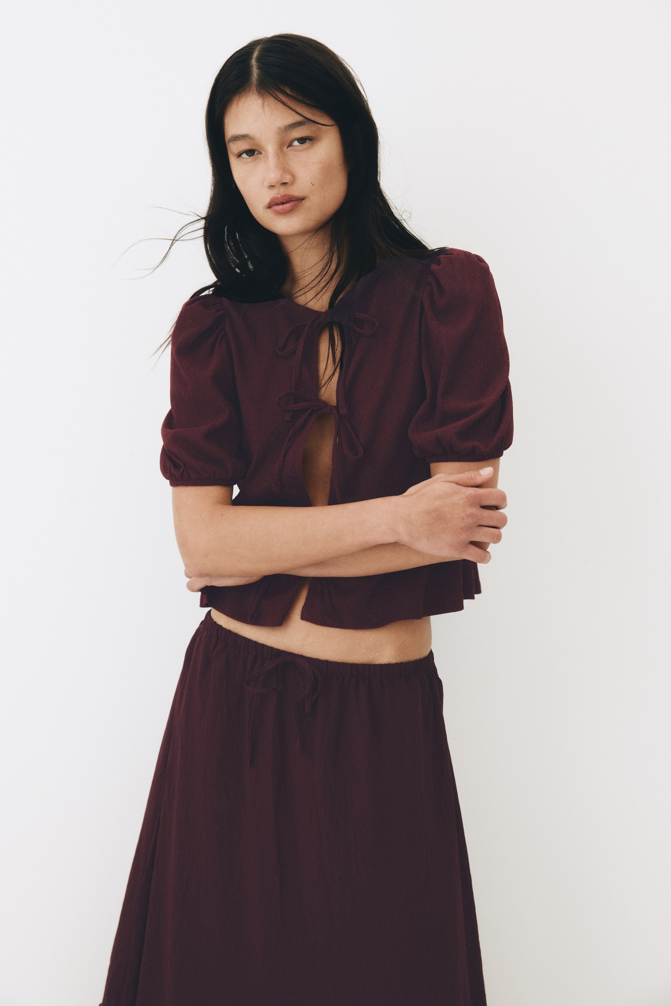 View larger image: The model is wearing a textured jersey top with a round neckline, featuring an open front secured by three sets of narrow ties. This top includes short puff sleeves with elasticated cuffs, a gathered waist seam, and a flared peplum. It is paired with a matching textured skirt, while the model poses with her arms crossed.