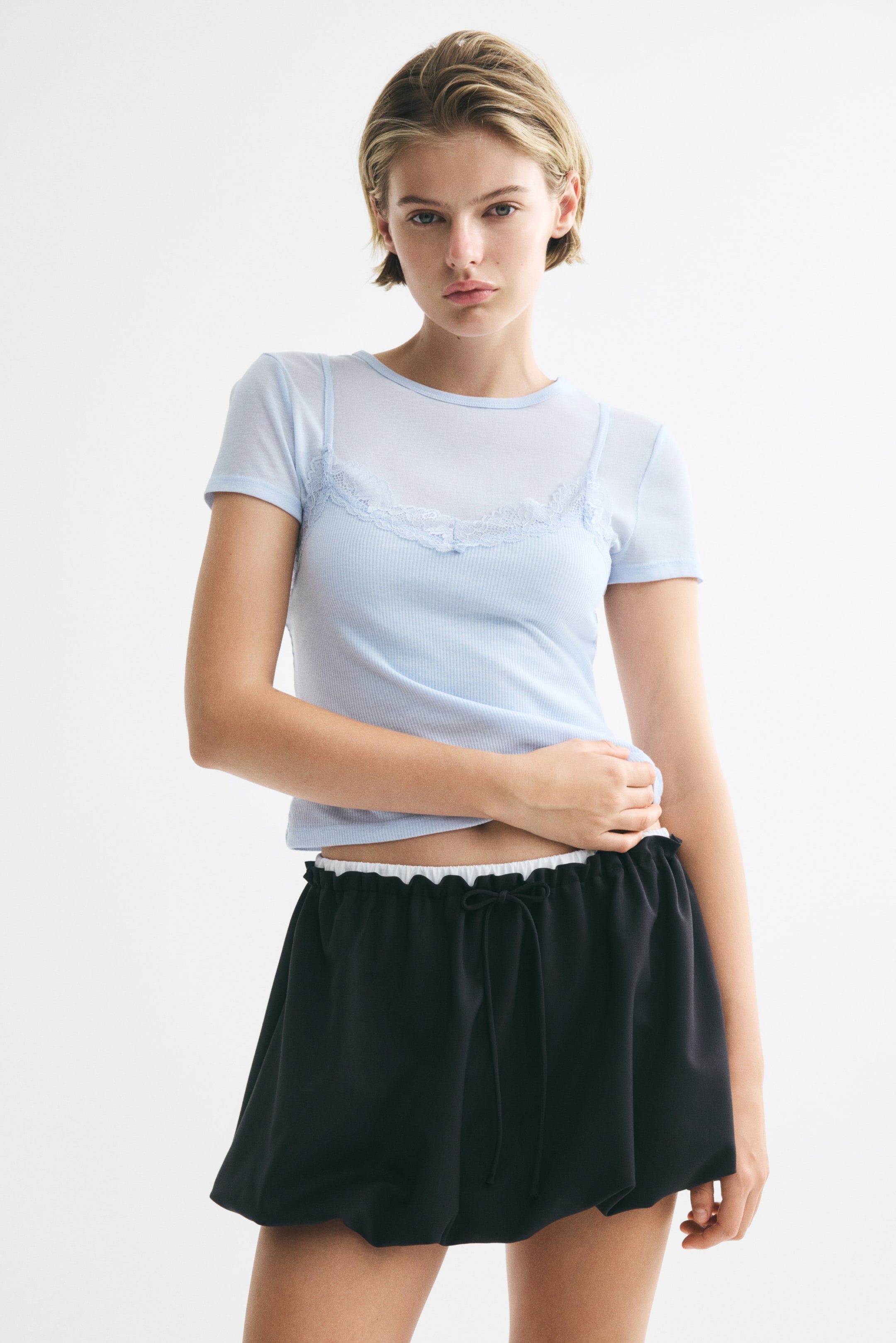 View larger image: A layered top in soft jersey is worn by the model, featuring an underlayer with a round neckline and short sleeves, and an outer layer with a V-neckline, scalloped lace trim, narrow shoulder straps, and a straight-cut hem. The model is gently holding the lower hem of the top.