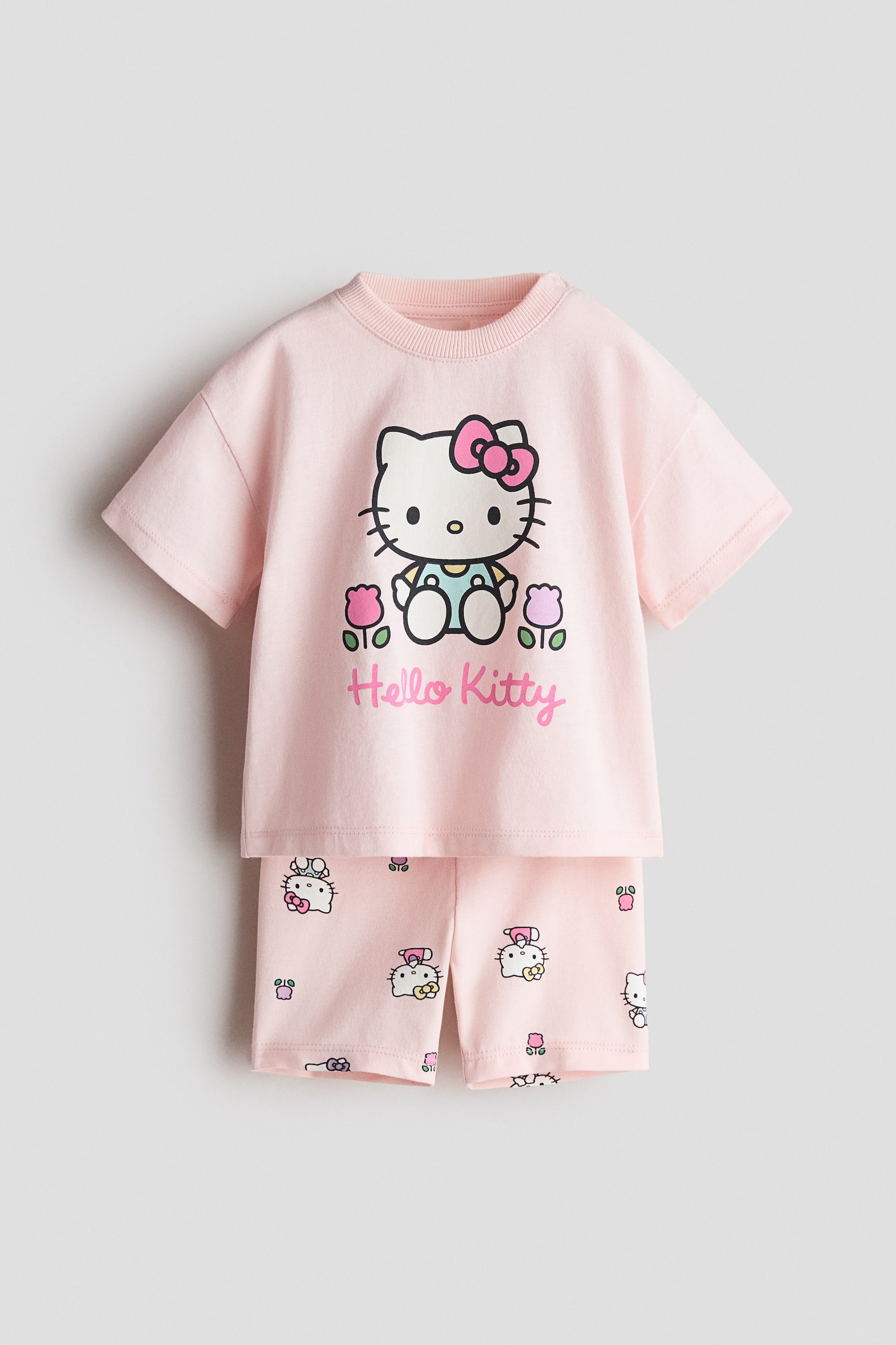 Voir une image plus grande: Un ensemble en coton rose de 2 pièces est présenté de face. Le haut à manches courtes présente un col rond côtelé et un grand imprimé graphique de Hello Kitty portant une salopette, entourée de tulipes, avec le texte "Hello Kitty" en dessous. Le short assorti arbore un motif intégral de petits visages de Hello Kitty et de fleurs.
