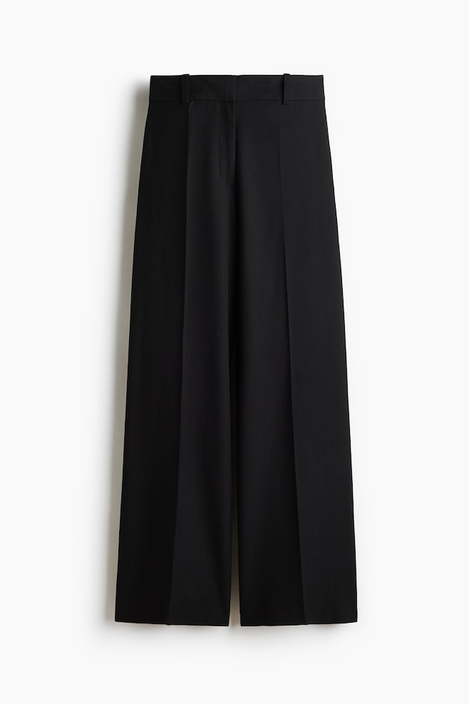 Front view of black tailored trousers in woven fabric, featuring a high waist, a visible zip fly, subtle side pockets, and wide legs with prominent creases running down the front. - 1