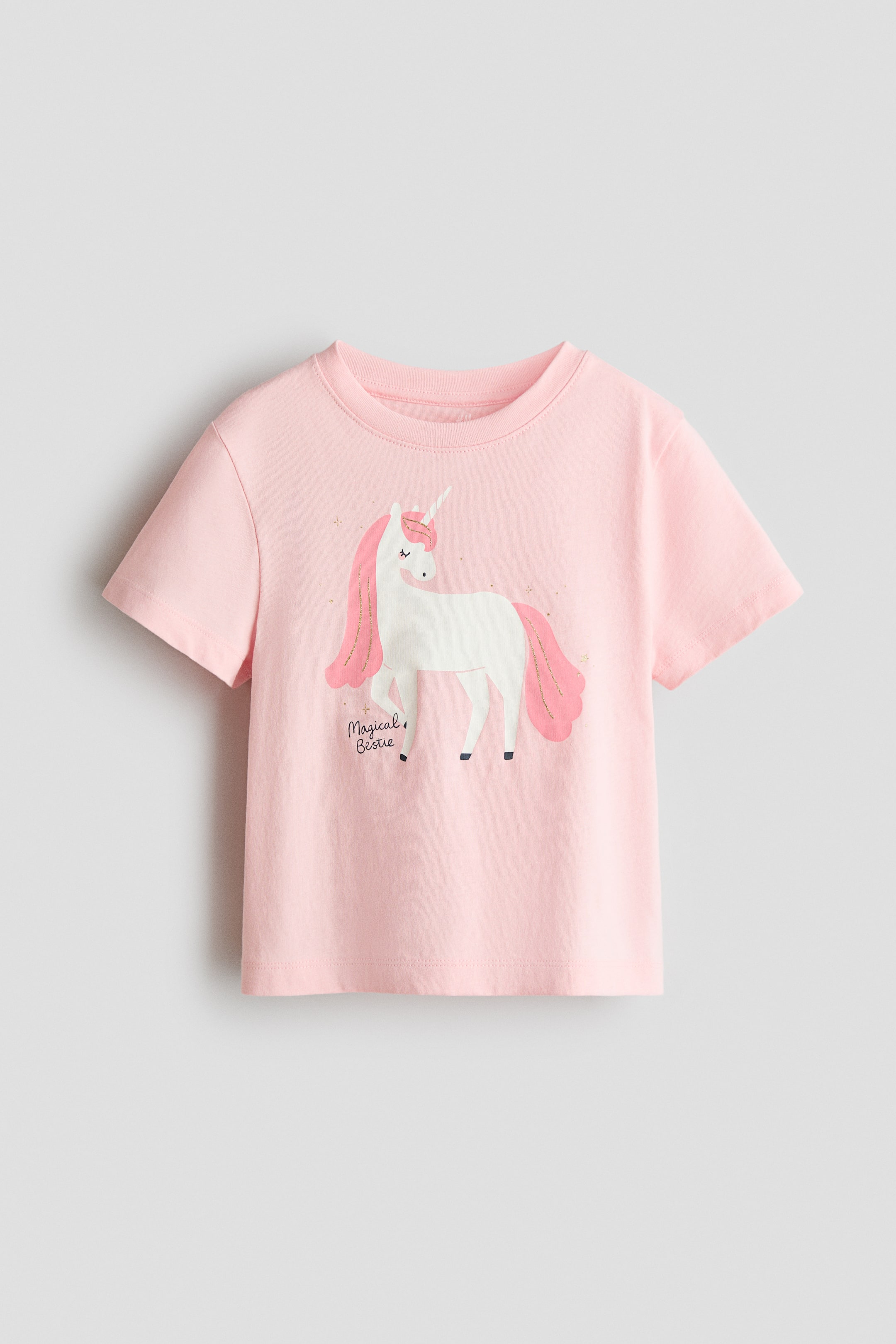 Xem ảnh lớn hơn: Áo thun hồng bằng vải cotton jersey in mềm mại với viền gân quanh cổ áo đang hướng về phía trước. Áo có hình in kỳ lân với bờm và đuôi màu hồng, cùng dòng chữ "Magical Bestie".