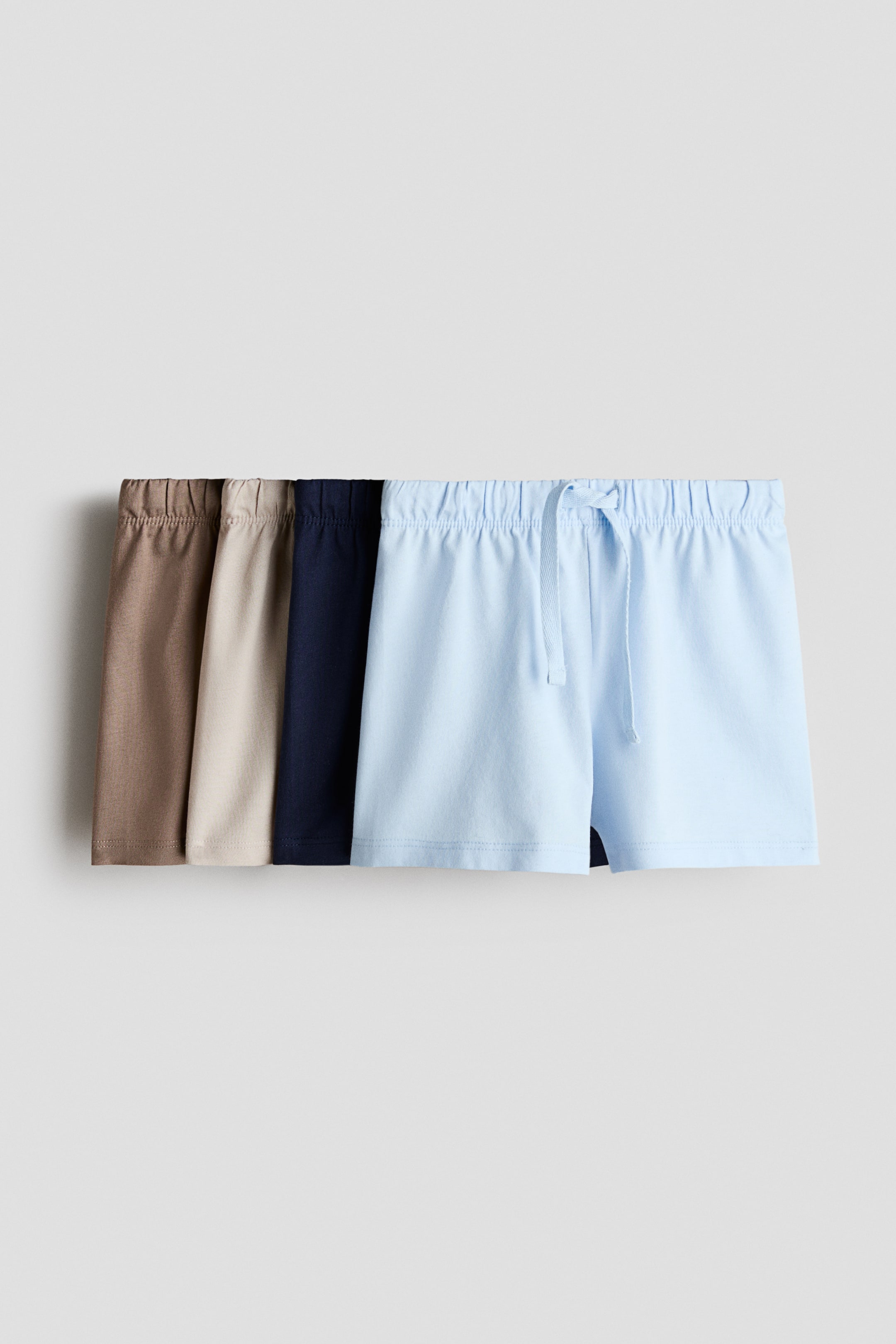 Visualizza immagine più grande: Un set di pantaloncini in morbido jersey di cotone è esposto, caratterizzato da vita elasticizzata. Sono visibili quattro paia nei colori marrone, beige, blu navy e azzurro chiaro, con il paio azzurro chiaro rivolto in avanti che mostra un laccio in vita.