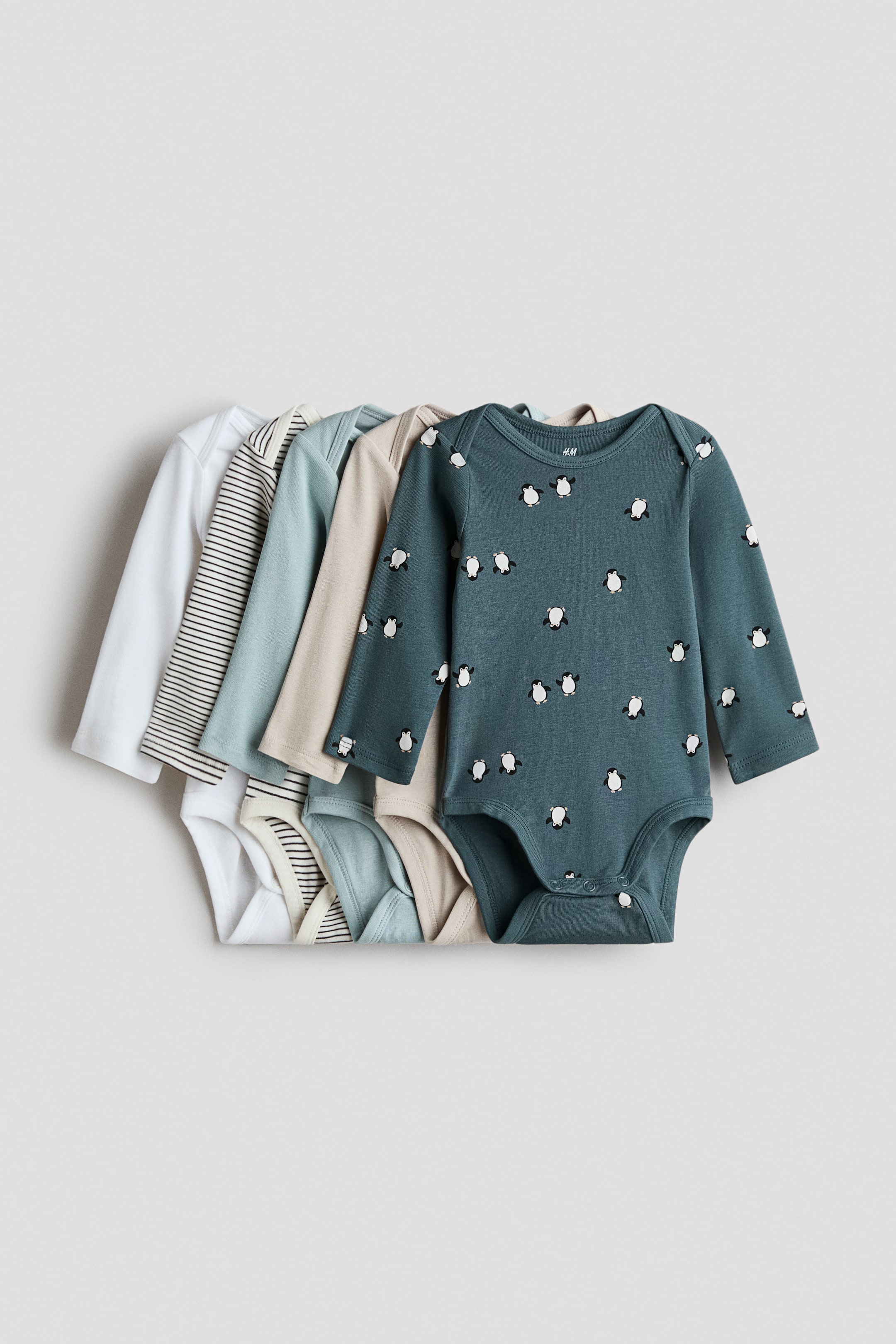 View larger image: Five long-sleeve baby bodysuits are arranged in an overlapping row, showcasing their front designs with envelope necklines and press-stud closures at the crotch. The assortment features a range of colours and patterns, including solid white, black-and-white stripes, light sage green, light beige, and a dark teal bodysuit decorated with a repeating penguin print.