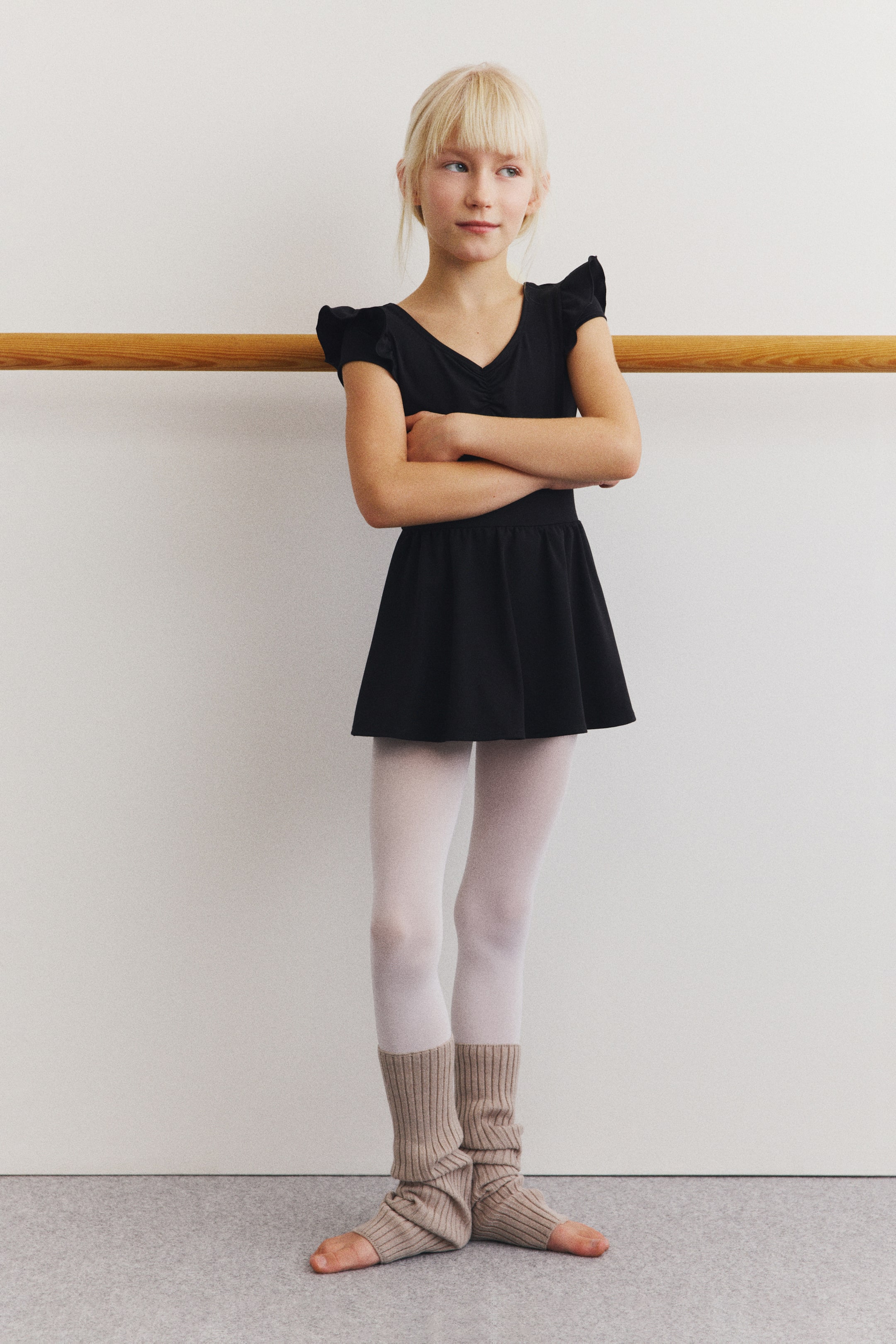 Vezi imaginea mai mare: Un leotard de dans negru, cu fustă atașată, este purtat și este prezentat din față. Prezintă un decolteu în V, mâneci scurte cu volane și o fustă atașată care se evazează din talie.