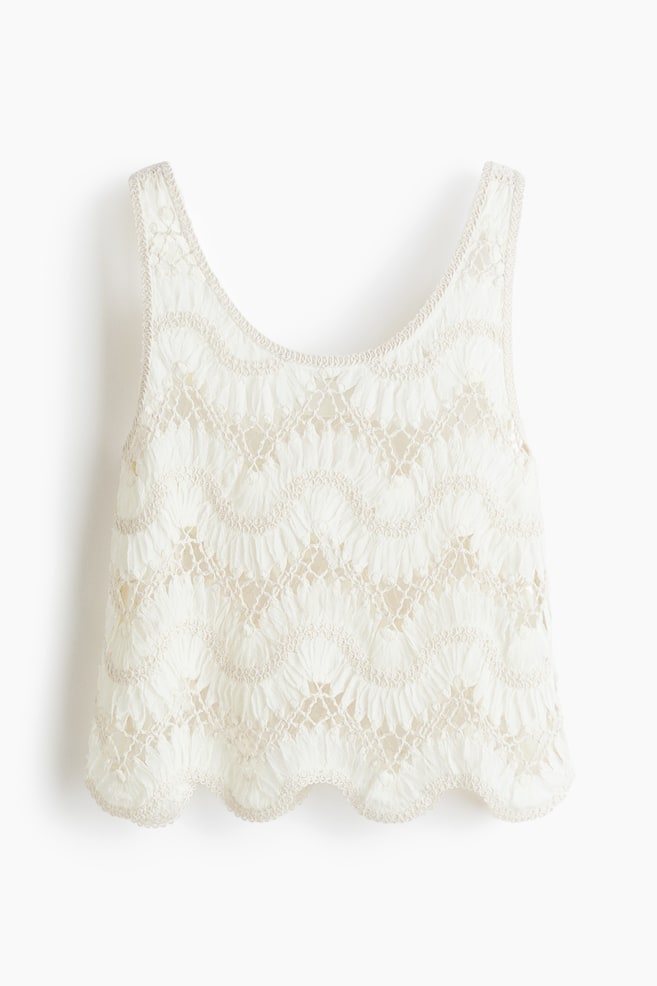 A cream-coloured sleeveless top is showcased from the front, featuring an intricate openwork lace pattern. The lace forms wavy, scalloped lines across the body, with a contrasting trim bordering the scoop neckline, armholes, and the scalloped bottom hem. - 1