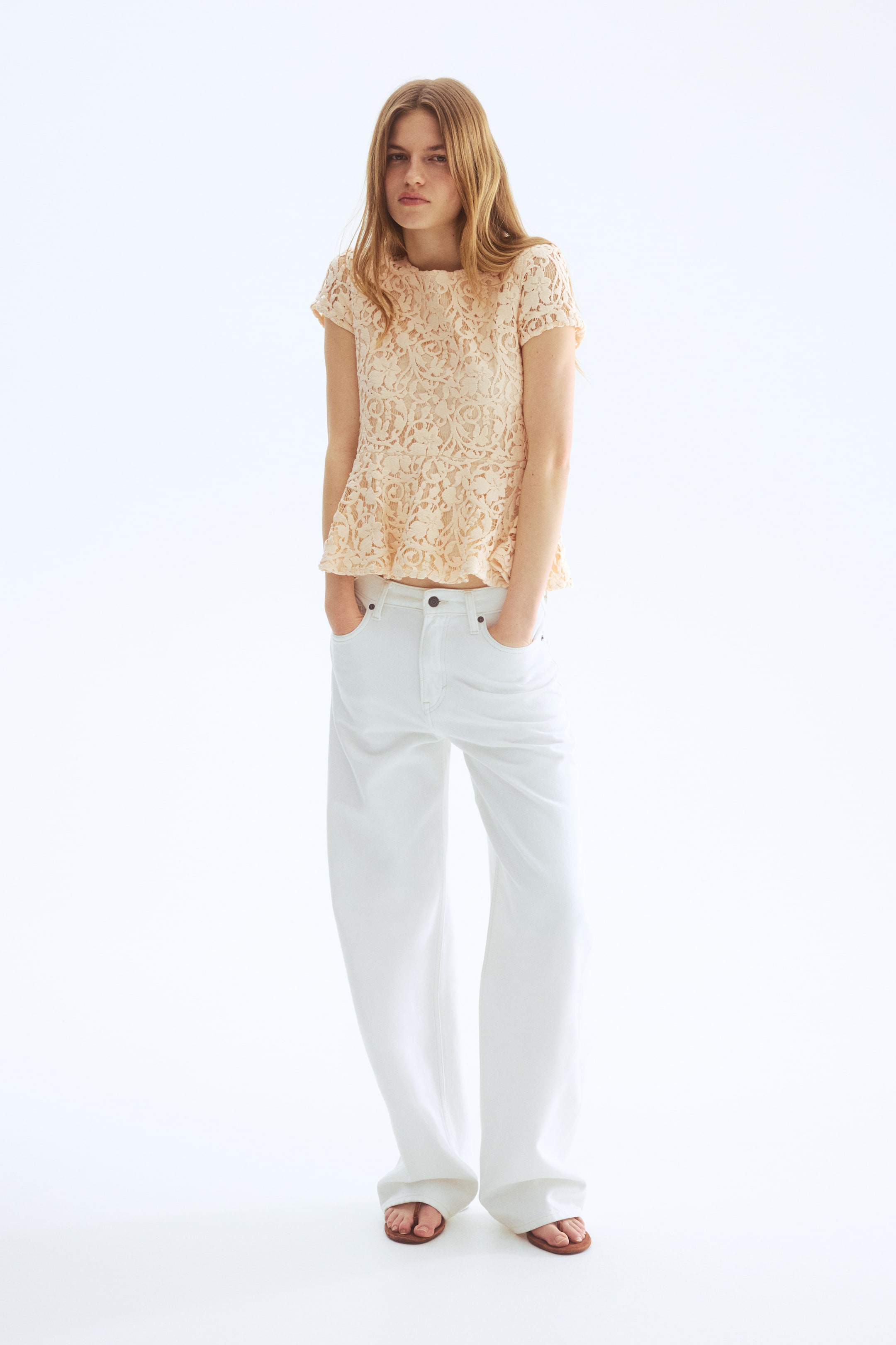 View larger image: The model is wearing a peach crochet-look knit top, showcasing a round neckline, cap sleeves, and a flared peplum. She pairs it with white wide-leg trousers and brown flat sandals.