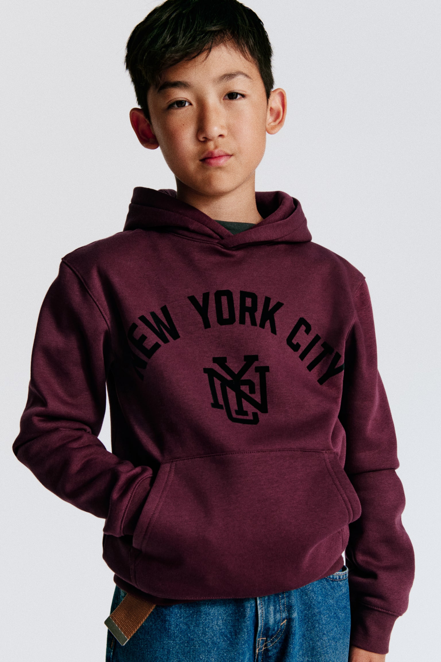 A dark red hoodie is showcasing a black "NEW YORK CITY" print with an "NYC" monogram on the front. It is crafted with a lined hood featuring a wrapover front, dropped shoulders, a kangaroo pocket, and ribbed cuffs and hem. - 1