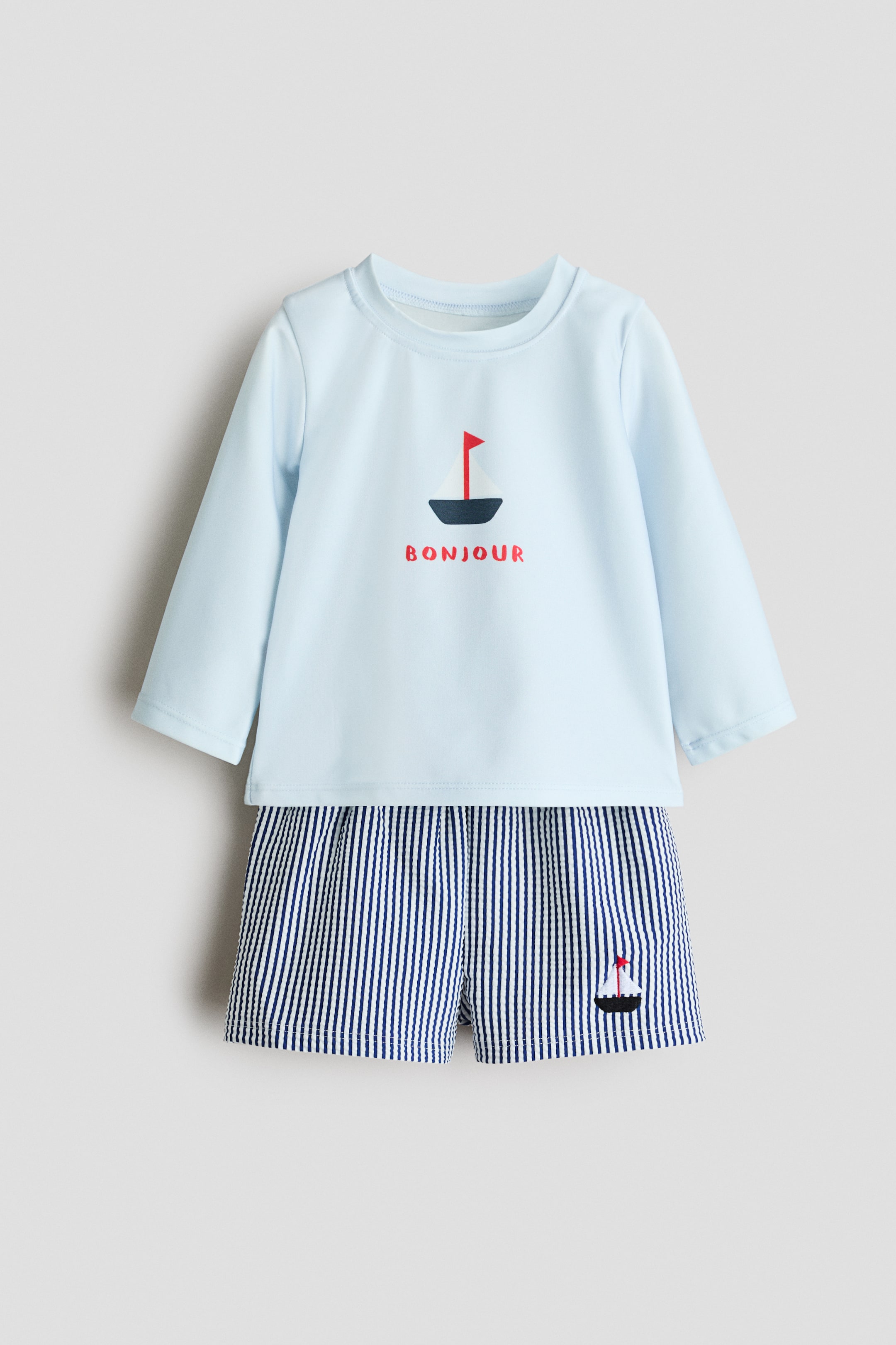 View larger image: A baby's two-piece outfit is laid flat, facing forward, featuring a light blue long-sleeved top paired with navy blue and white vertical striped shorts. The top showcases a sailboat graphic with a red flag and the word 'BONJOUR' on the front, while the shorts sport a small sailboat graphic on the right leg.
