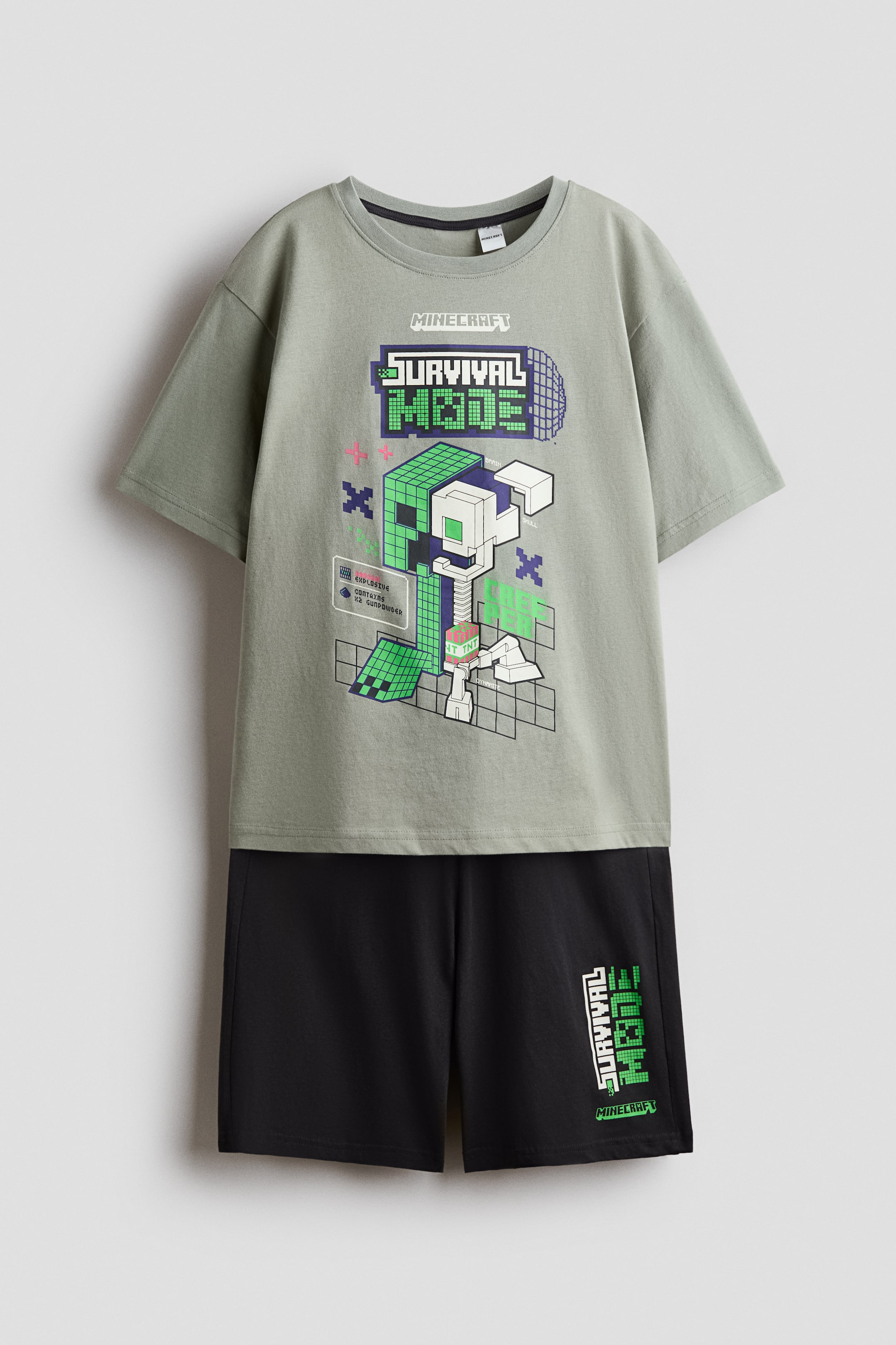 View larger image: A two-piece set featuring a light olive green, short-sleeved t-shirt with a crew neck and a pixelated "MINECRAFT SURVIVAL MODE" graphic showcasing a green Creeper and a white skeleton on the front. The accompanying black shorts display "SURVIVAL MODE" text vertically in green pixelated font on the left leg.