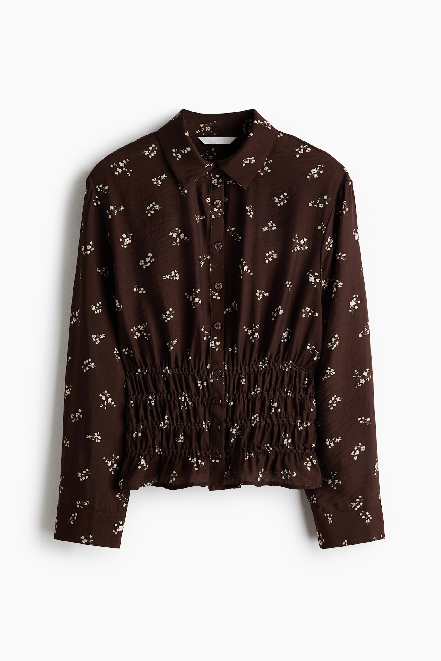 A dark brown blouse featuring a small white floral pattern is shown from the front, displaying a shirt collar, a button-down front, and long sleeves with buttoned cuffs. The blouse showcases a smocked, elasticated waist that creates a gathered, defined silhouette with a slightly ruffled hem. - 1