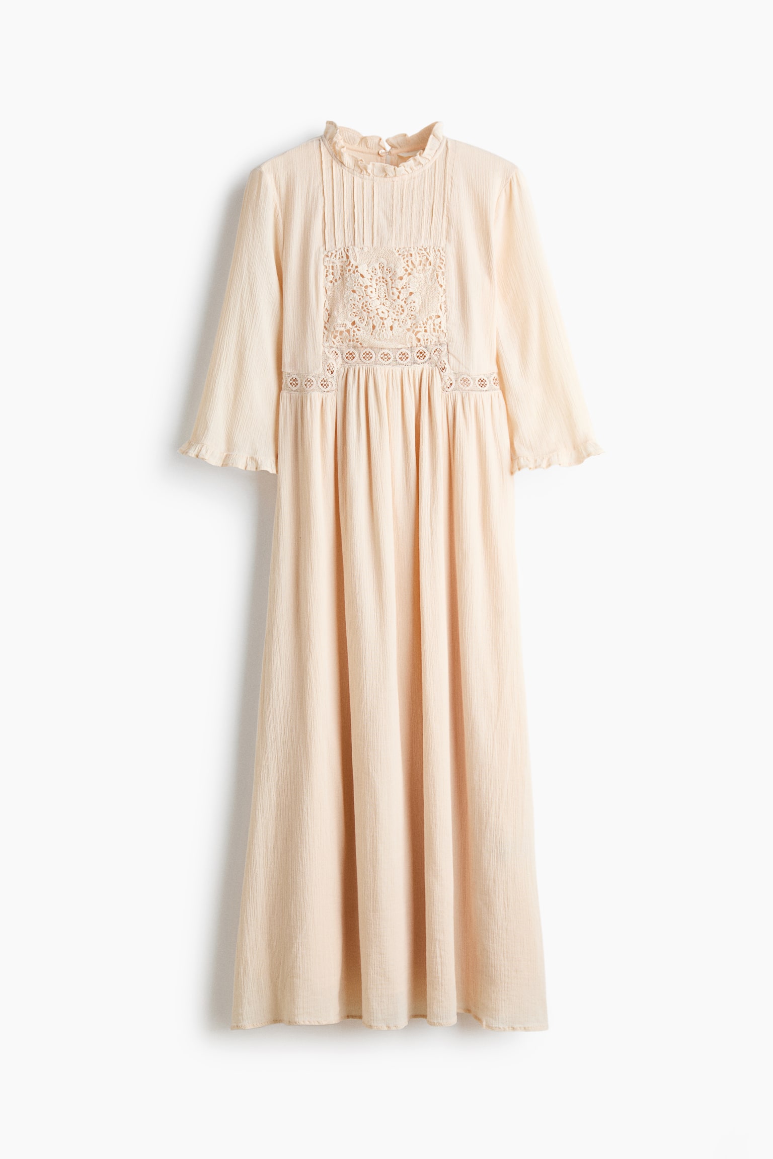 A long, light beige dress in a crinkled cotton weave is shown front-facing, featuring a high neckline with frill trim, vertical pintucks, and a lace inset panel on the bodice. The design includes three-quarter-length sleeves with ruffled cuffs and a gathered waist that flows into a long, gently flared skirt.