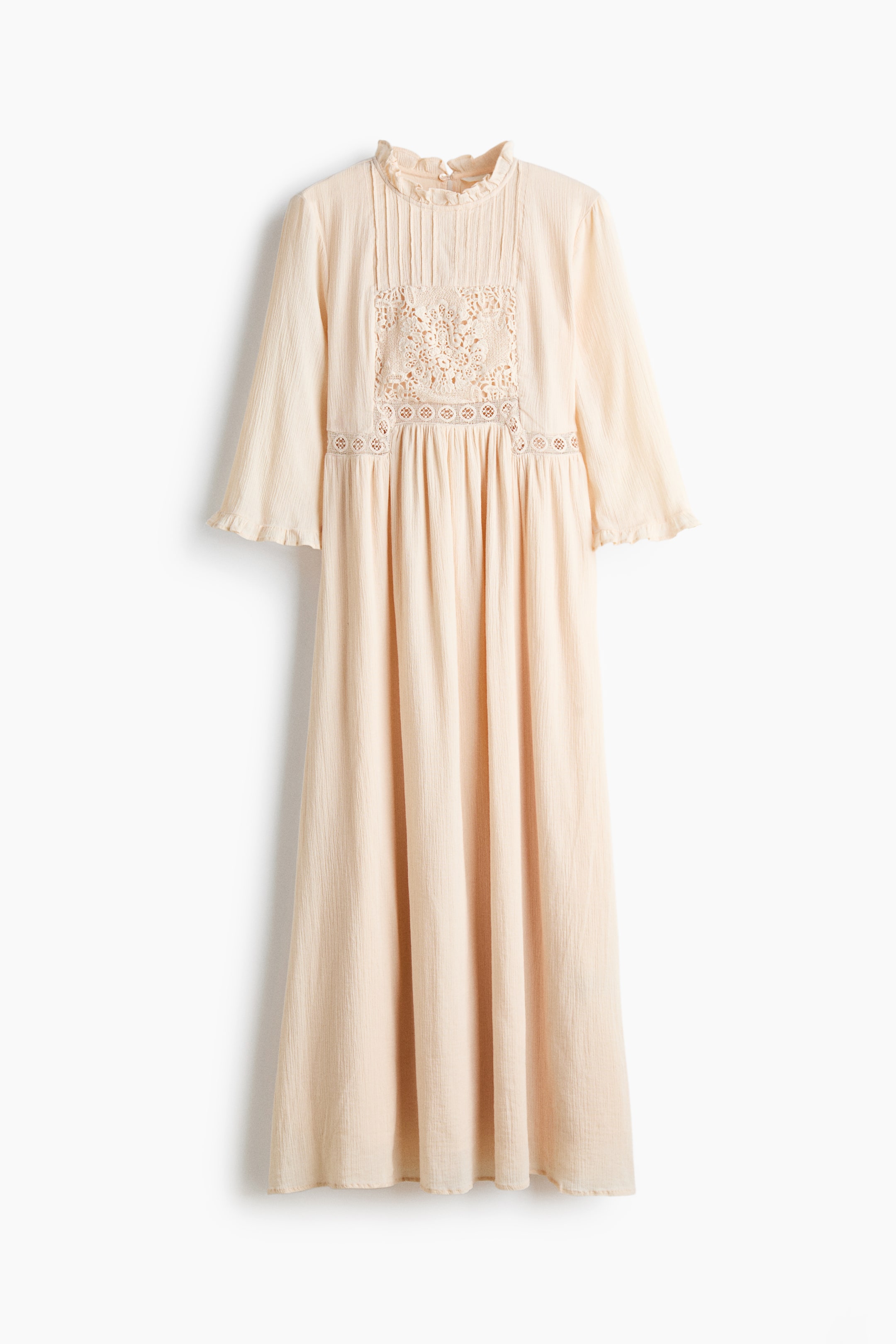 View larger image: A long, light beige dress in a crinkled cotton weave is shown front-facing, featuring a high neckline with frill trim, vertical pintucks, and a lace inset panel on the bodice. The design includes three-quarter-length sleeves with ruffled cuffs and a gathered waist that flows into a long, gently flared skirt.