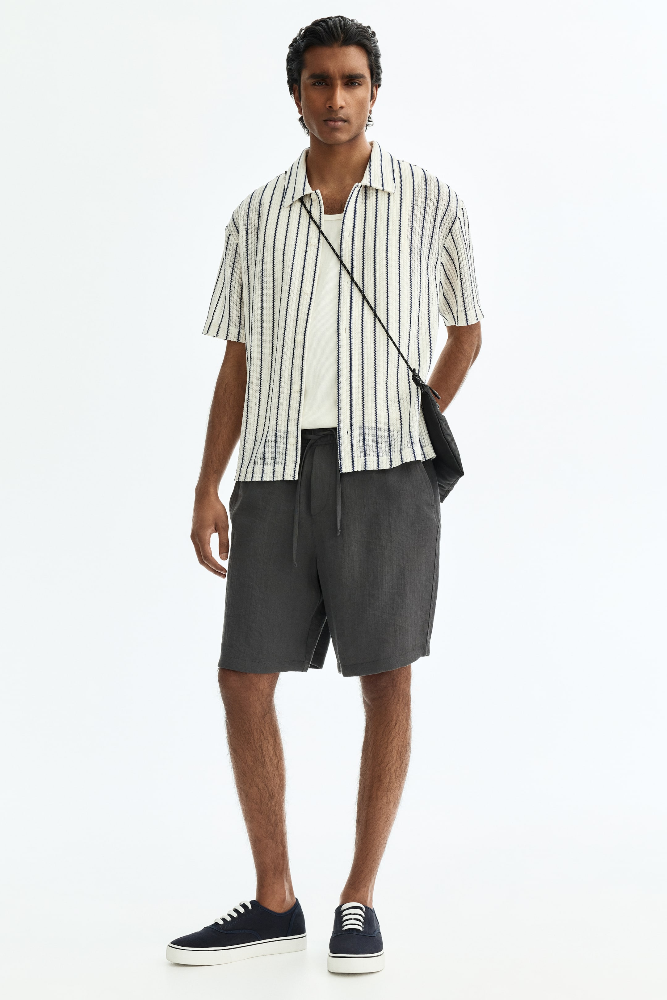 Ampliar la imagen: El conjunto se presenta de frente, con una camisa color crema de manga corta con cuello y rayas verticales texturizadas en azul oscuro, llevada abierta sobre una camiseta lisa de cuello redondo color crema. Unos shorts gris oscuro con cordón y cintura ajustable, zapatillas de lona azul marino con cordones blancos, y un pequeño bolso negro cruzado completan el look.