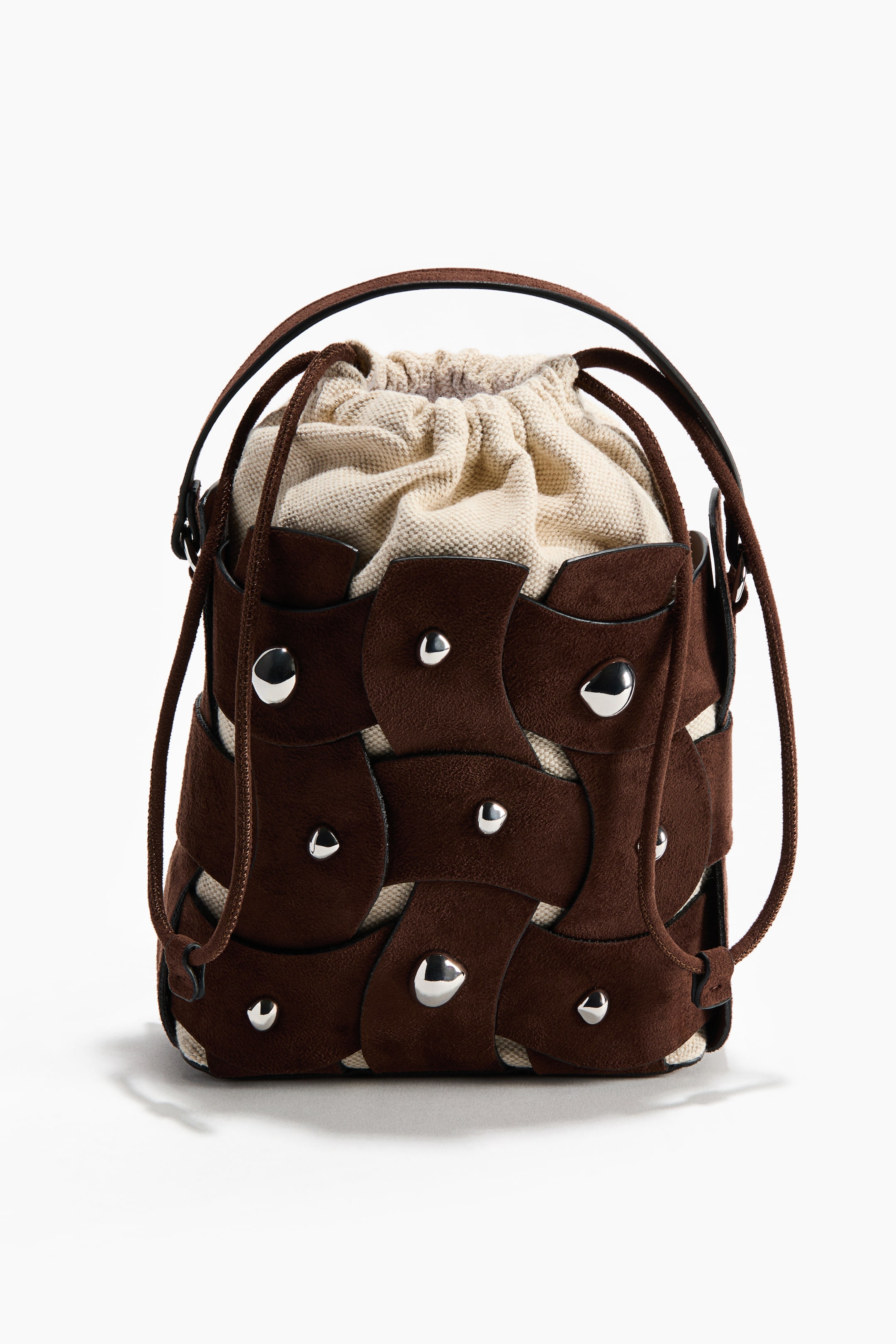 View larger image: A brown bucket bag is shown from the front, featuring dark-brown interlocking suede-like panels trimmed with silver-toned studs. A light-coloured canvas drawstring pouch is visible inside, along with slim dark-brown shoulder straps and a matching drawstring cord.
