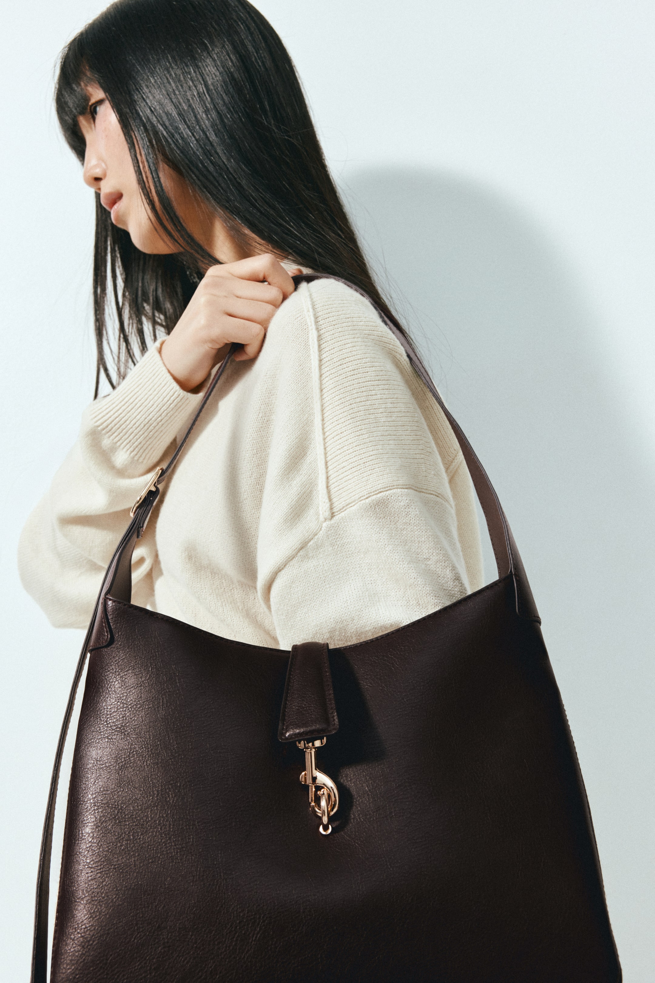 View larger image: A dark brown shoulder bag in textured coated fabric is worn over the shoulder by the model, who is holding the strap with one hand. The bag features an adjustable strap with a metal buckle and a front tab with a gold-toned carabiner hook clasp.