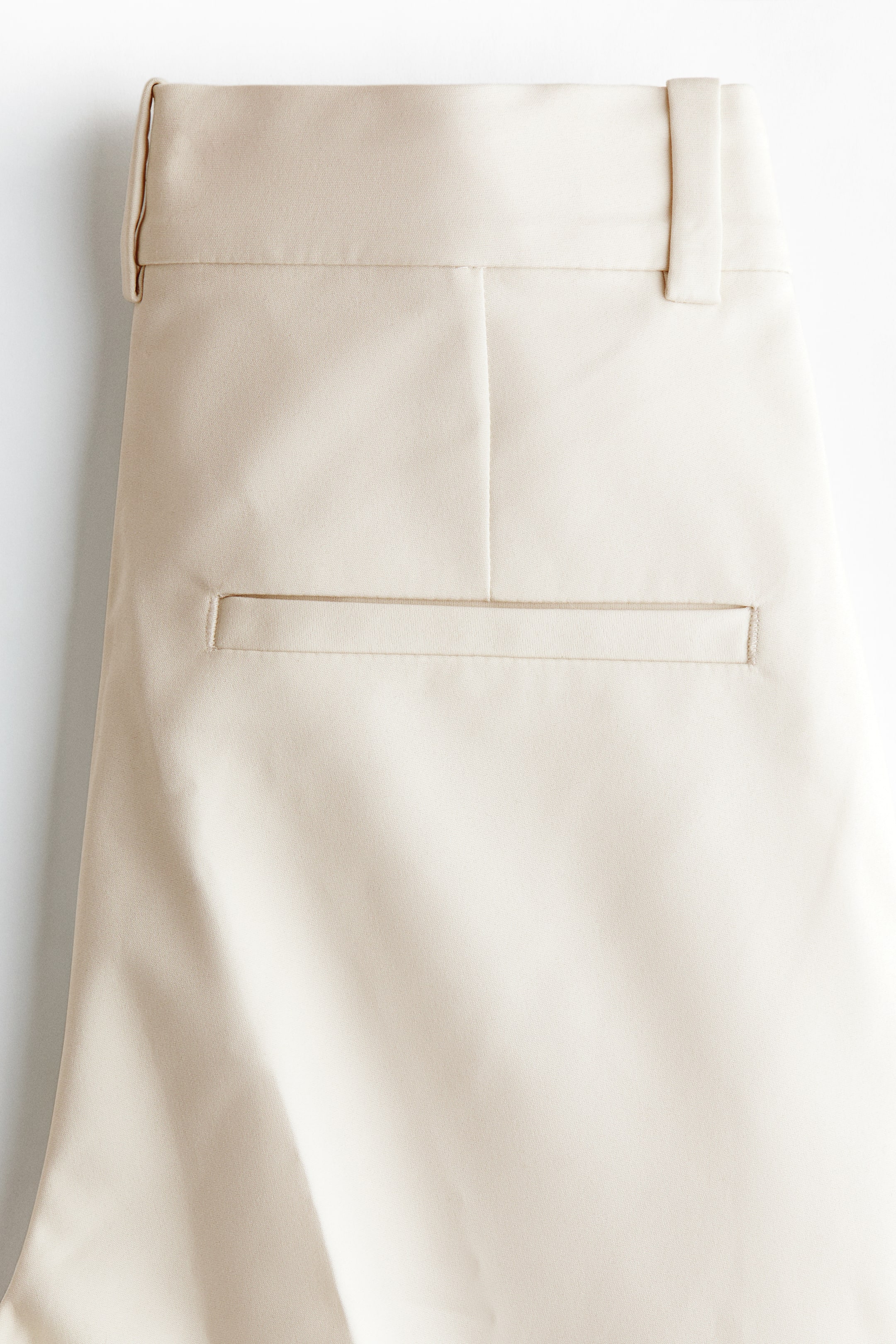 Ampliar la imagen: Pantalones cigarrete de largo al tobillo, confeccionados en un tejido elástico beige claro, muestran su vista trasera, con una pretina regular, una trabilla visible y un falso bolsillo trasero de vivo horizontal.