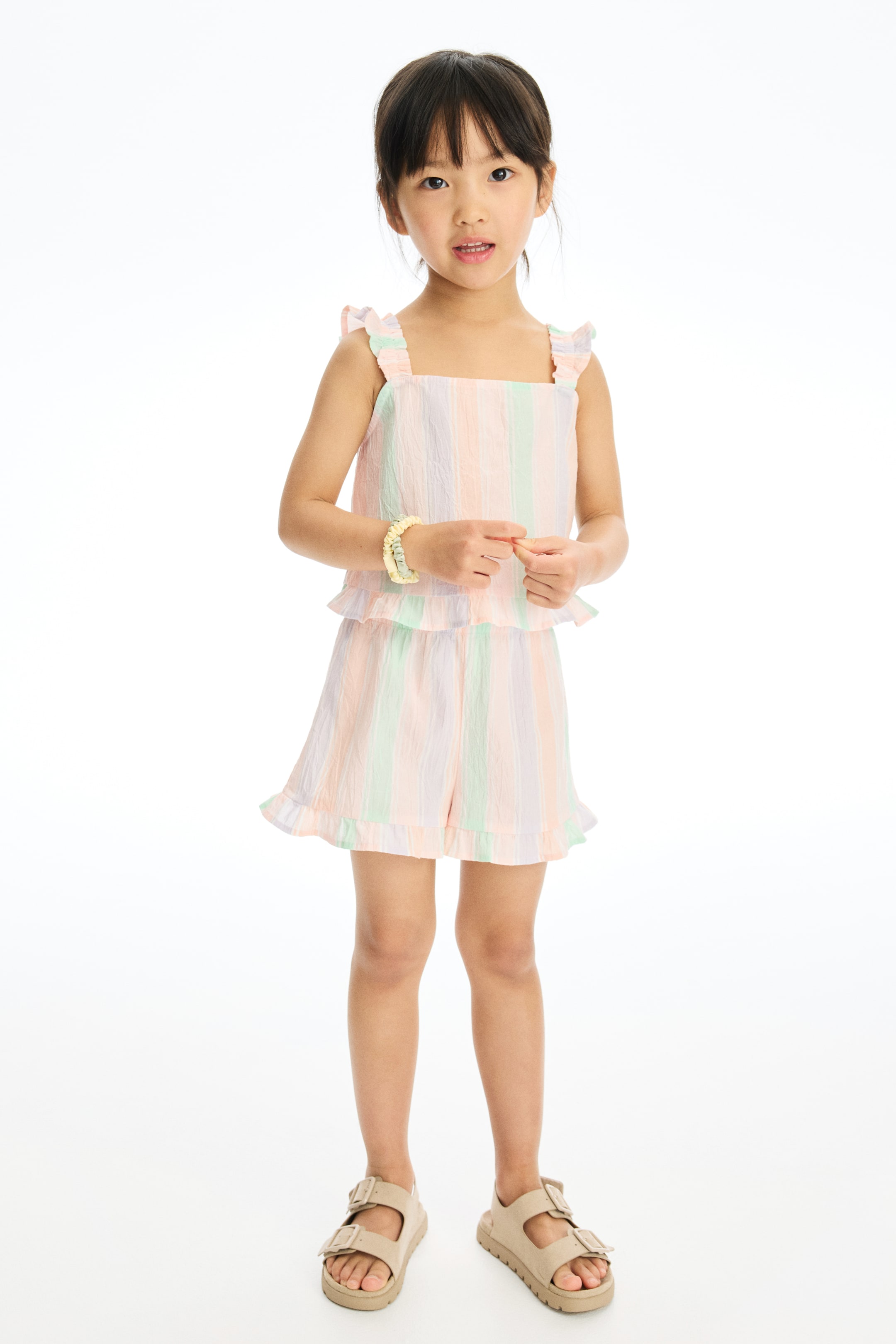 View larger image: A two-piece child's outfit featuring a square-neck top and matching shorts, presented front-facing. The top is designed with ruffled shoulder straps and a peplum hem, and the shorts showcase ruffled leg openings; both pieces display vertical pastel stripes in pink, green, and lavender with a crinkled texture.