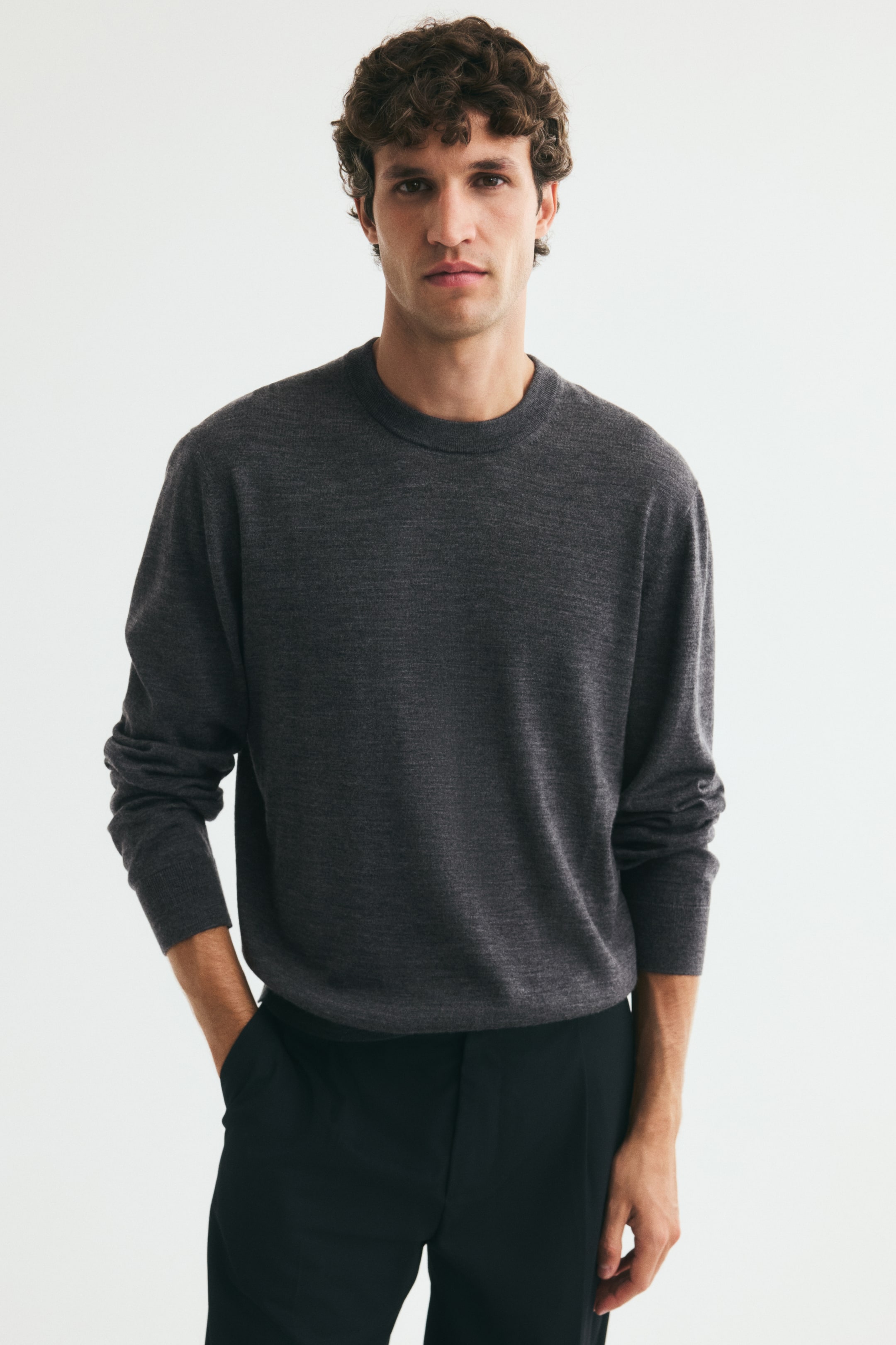 View larger image: A dark grey, fine-knit jumper featuring a round, rib-trimmed neckline and ribbed cuffs is worn by the model. The regular-fit jumper is paired with black trousers, with the model resting one hand in a pocket.