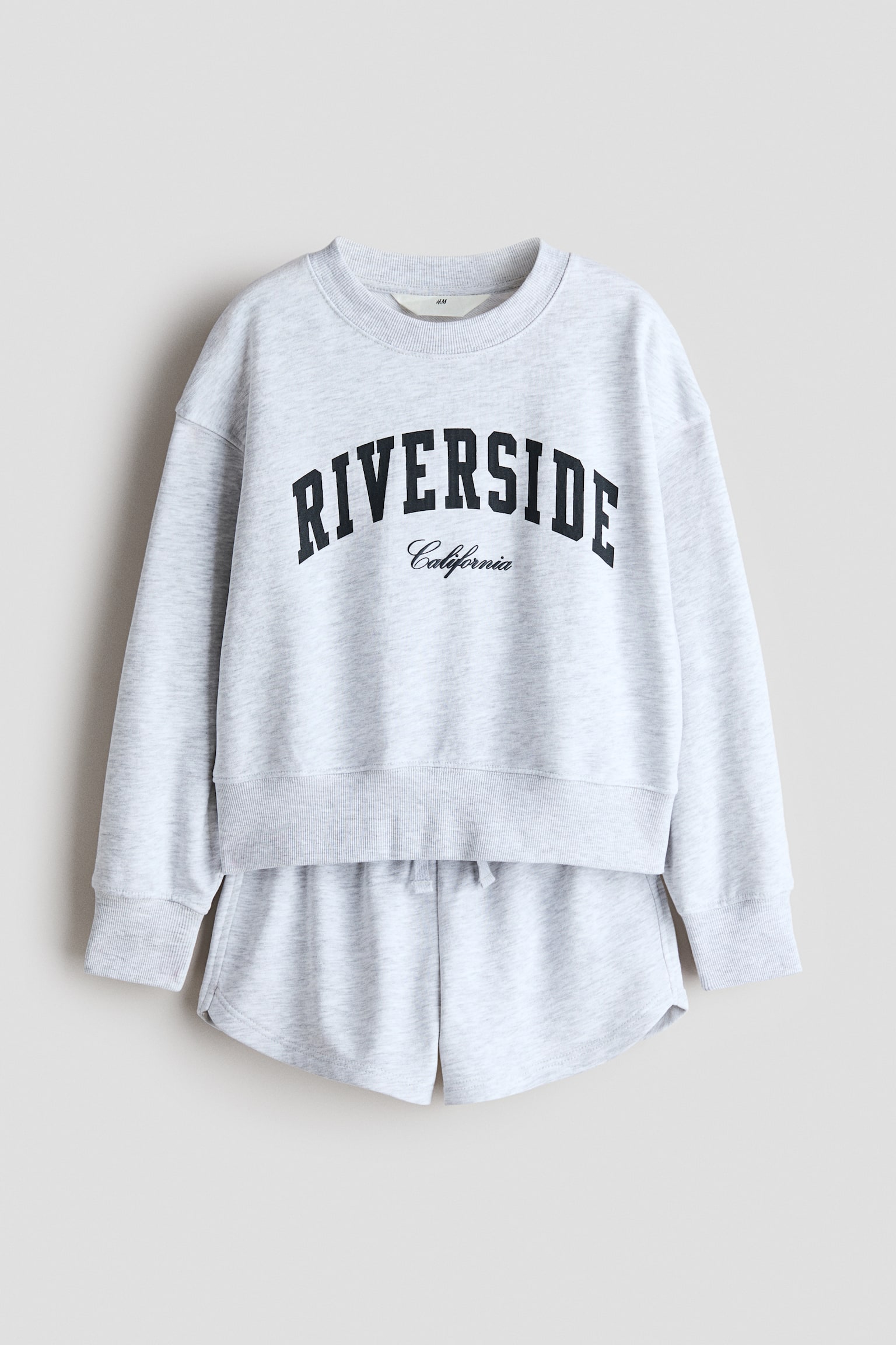 A light grey heathered sweatshirt and sweatshorts set is shown from the front. The sweatshirt features a ribbed round neckline, dropped shoulders, long sleeves with wide ribbed cuffs, and a black 'RIVERSIDE California' graphic across the chest. The matching shorts feature an elasticised waistband with a drawstring.