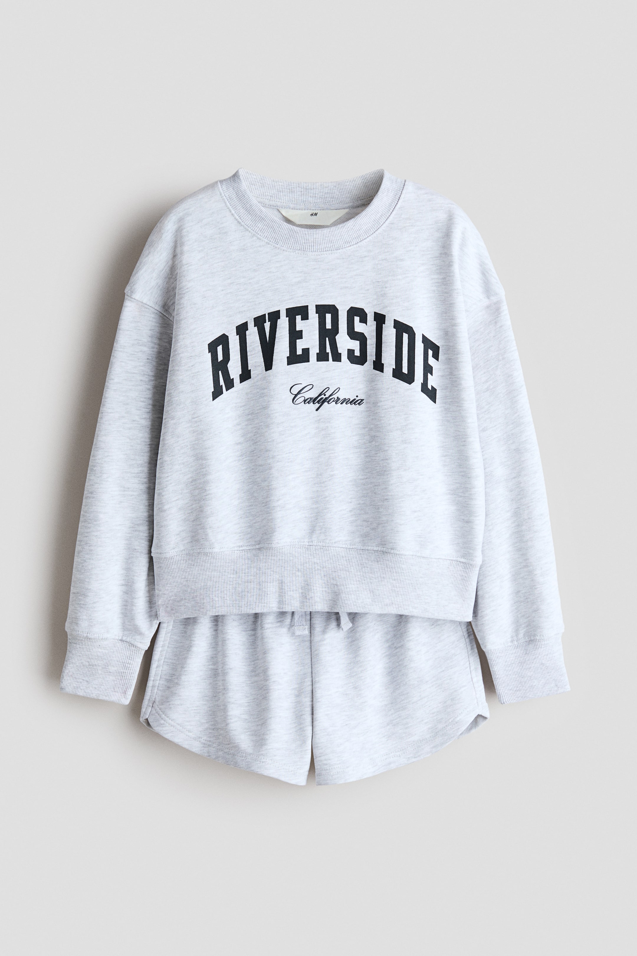 View larger image: A light grey heathered sweatshirt and sweatshorts set is shown from the front. The sweatshirt features a ribbed round neckline, dropped shoulders, long sleeves with wide ribbed cuffs, and a black 'RIVERSIDE California' graphic across the chest. The matching shorts feature an elasticised waistband with a drawstring.