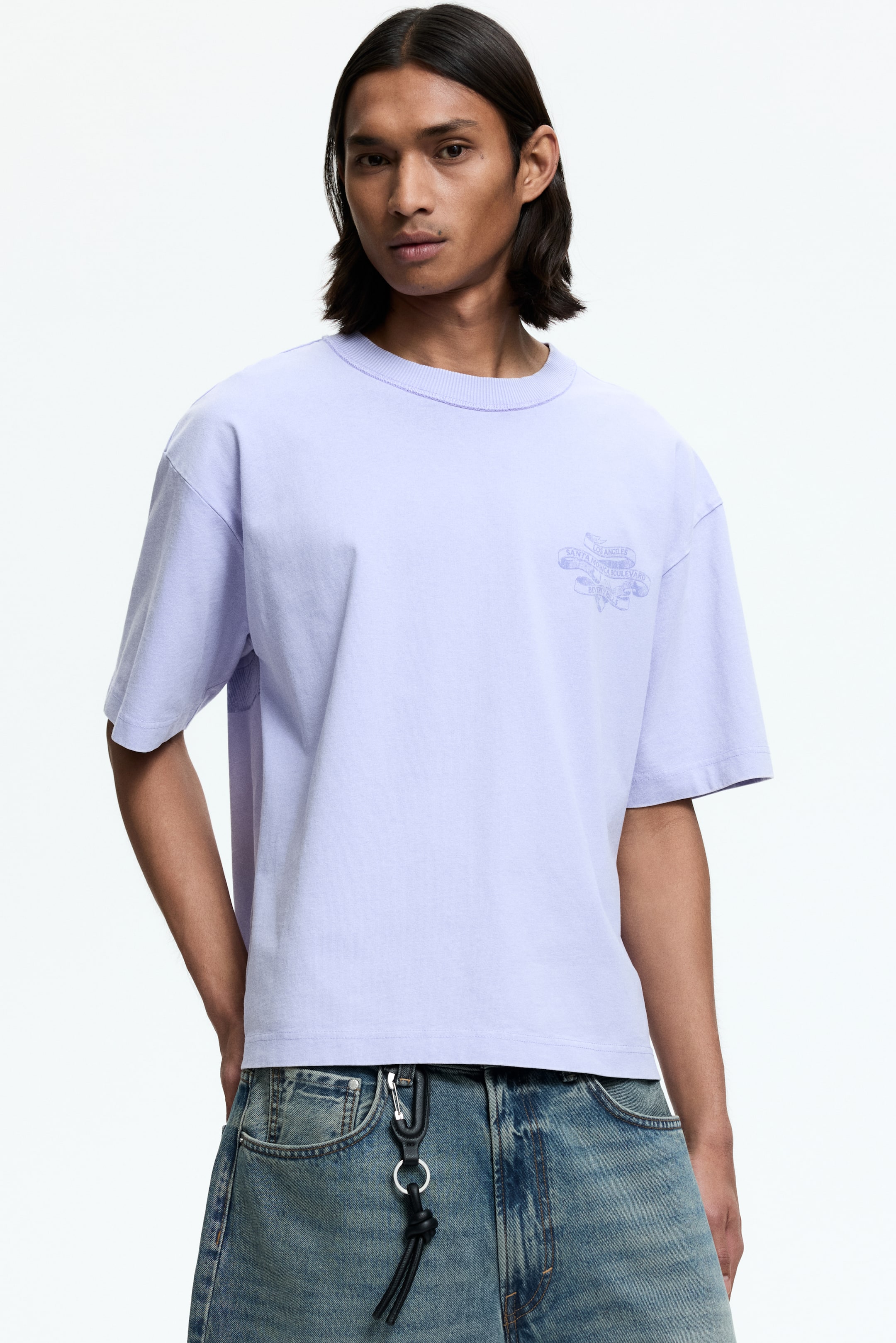 Ver imagen más grande: Una camiseta de color púrpura claro presenta un cuello redondo acanalado y mangas cortas y holgadas. Un gráfico sutil en un tono púrpura más claro aparece en el pecho izquierdo, con la camiseta mirando hacia adelante.