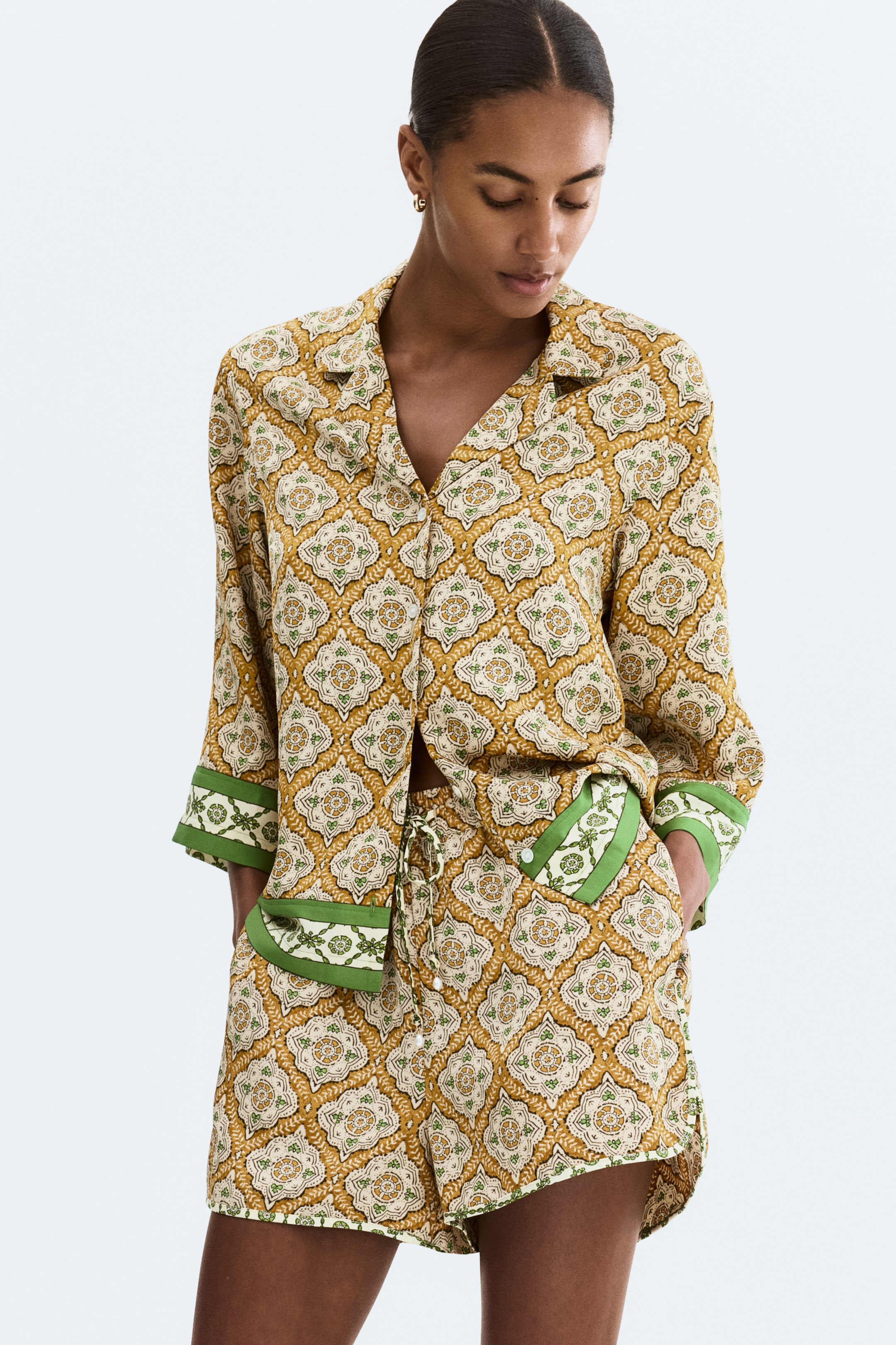View larger image: The product is a matching shirt and shorts set, featuring a mustard yellow, cream, and green geometric and floral pattern. The button-down shirt showcases a revere collar and rolled long sleeves with green patterned cuffs, while the shorts feature a drawstring waist and green-trimmed side pockets.