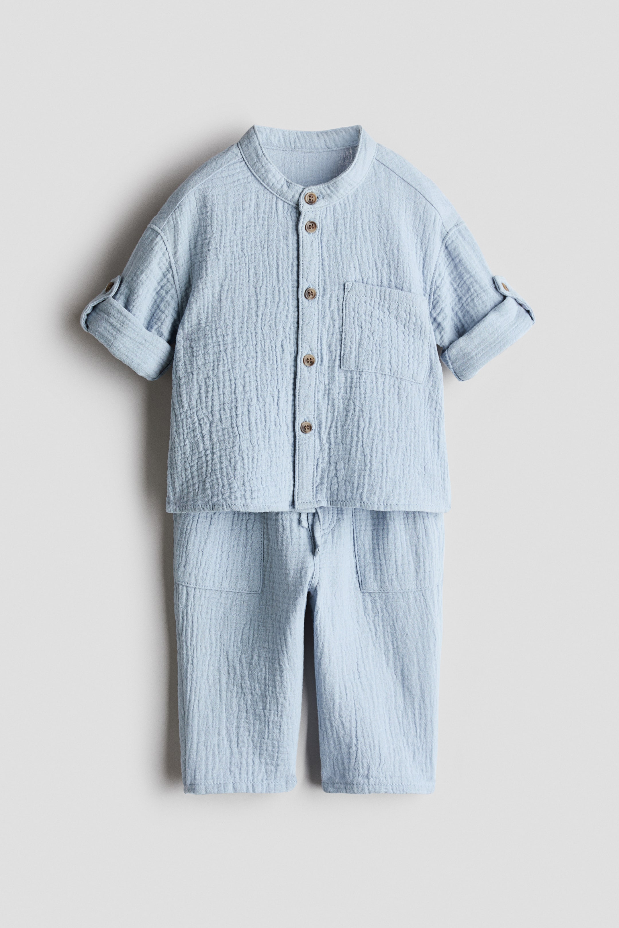 拡大画像を表示: ライトブルーのコットンモスリンセットは、シャツとマッチングトラウザーを特徴とし、正面を向いています。シャツはバンドカラーでデザインされ、前面には目立つブラウンのボタンが並び、オープンチェストポケットとタブとボタンで留められたロールアップスリーブが特徴です。トラウザーは、ゴム入りのドローストリングウエストとサイドポケットを備えています。