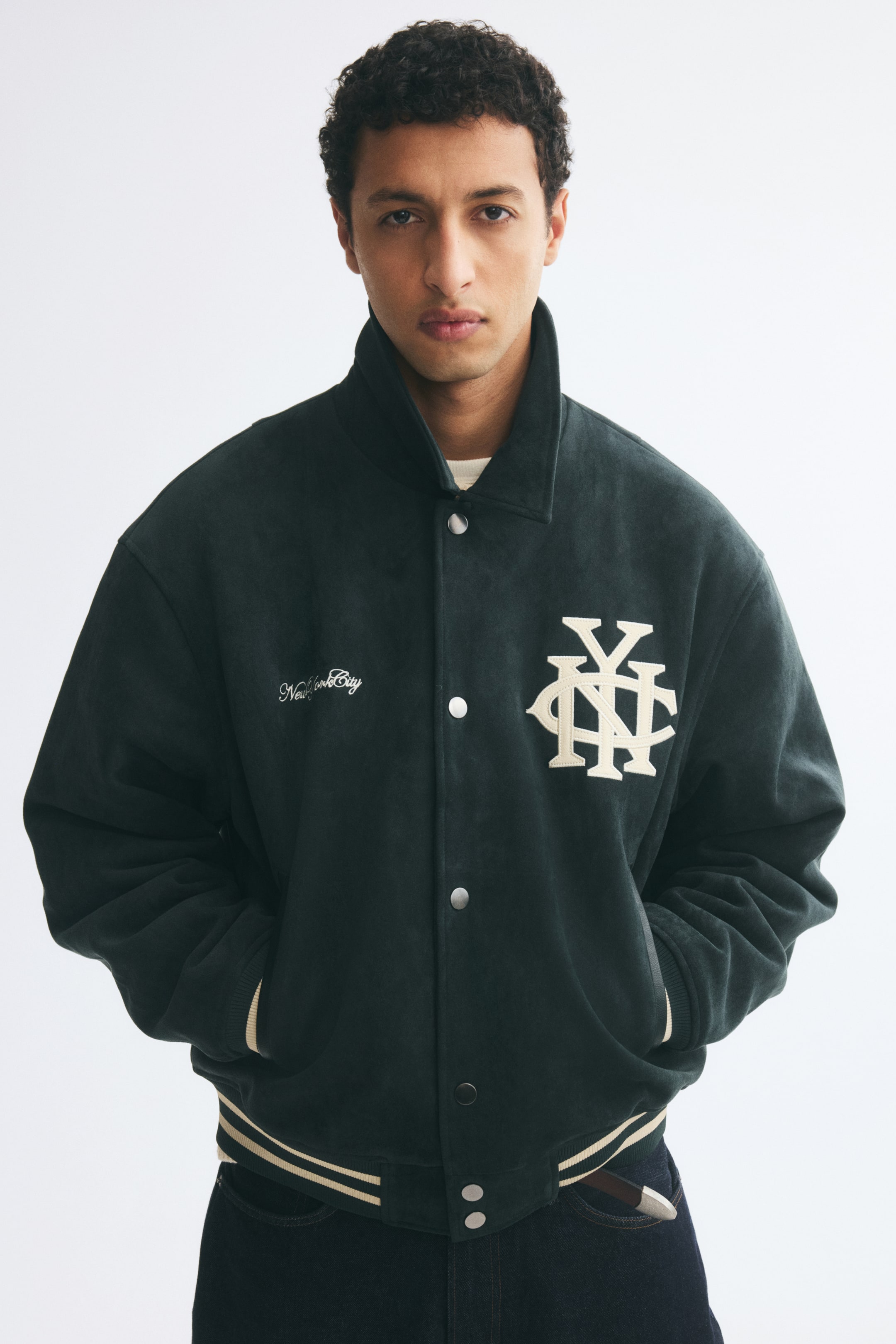 Größeres Bild anzeigen: Eine dunkelgrüne Baseballjacke aus angerautem Stoff wird vom Model getragen, das mit den Händen in den Eingrifftaschen steht. Die Jacke verfügt über einen Kragen, silberfarbene Druckknöpfe auf der Vorderseite, einen gestreiften Rippbund und cremefarbene Stickdetails auf der Brust, darunter ein großes Monogramm und Schriftzug.