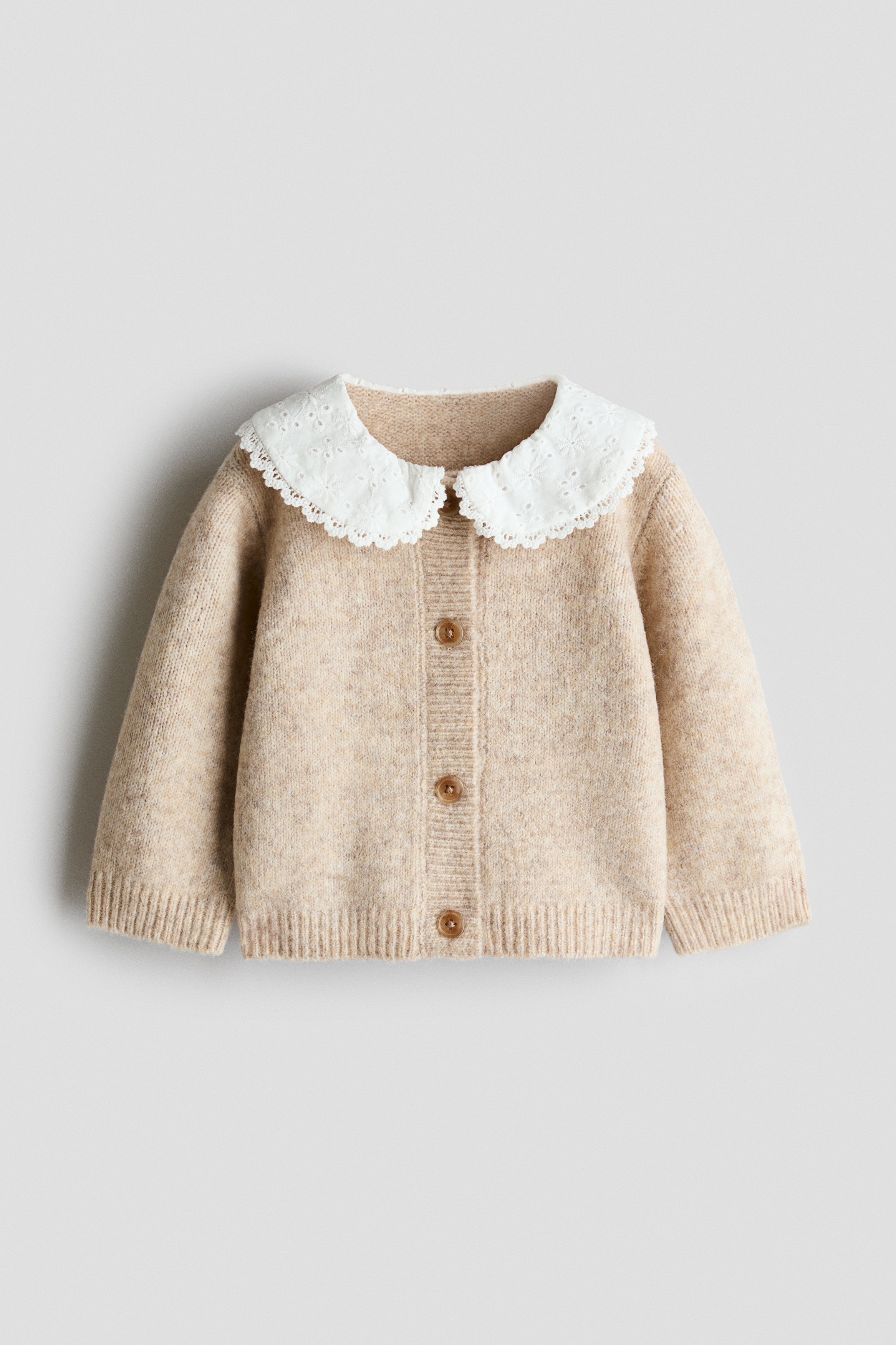 This light beige, fine-knit cardigan is shown from the front, featuring a prominent white rounded broderie anglaise collar with scalloped lace trim. The cardigan has brown buttons down the front and ribbed cuffs on the long sleeves.