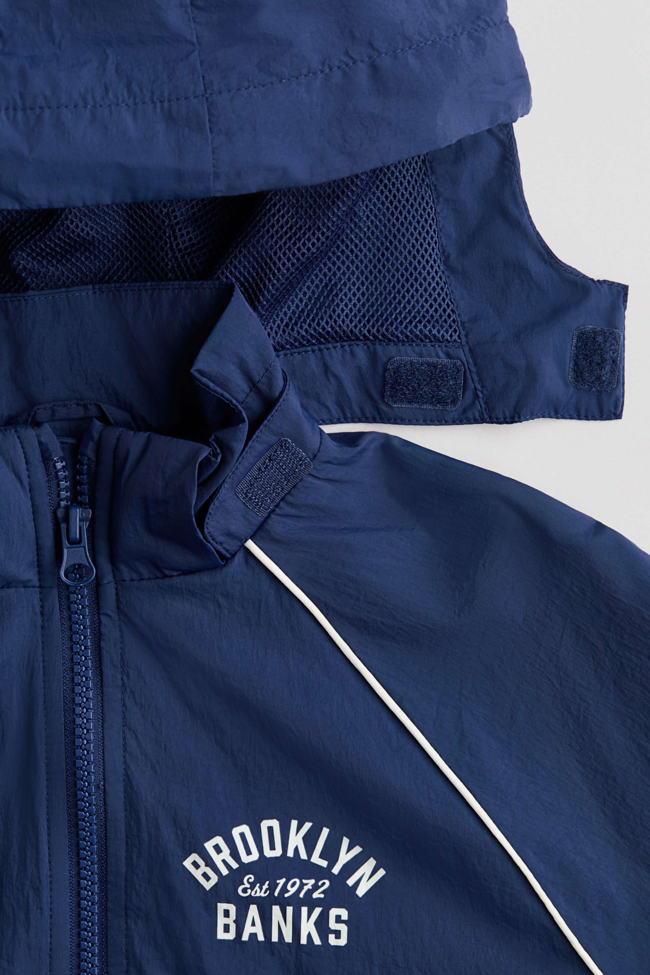 Visualizza l'immagine ingrandita: Una giacca blu navy presenta una cerniera a tutta lunghezza, un collo alto con patte in velcro e un cappuccio attaccato con fodera in mesh blu navy. Un piping bianco corre diagonalmente sul davanti della giacca, che riporta la scritta bianca "BROOKLYN Est 1972 BANKS."