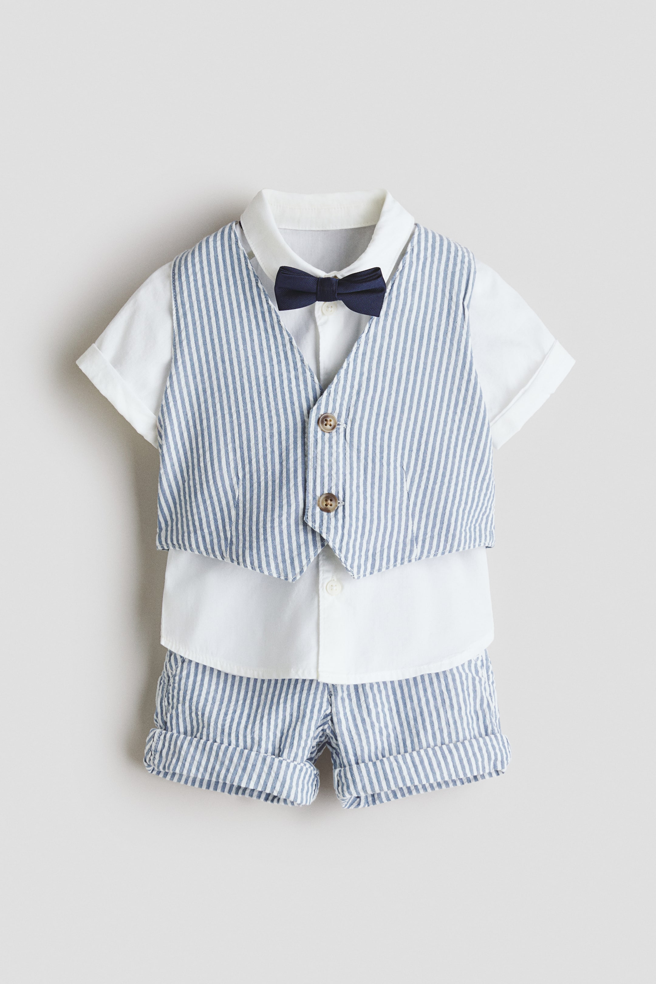 View larger image: A stylish ensemble is showcased from the front, featuring a blue and white vertically striped waistcoat with a soft, textured weave and two visible buttons, layered over a smooth white short-sleeved shirt. A navy blue bow tie embellishes the shirt, while matching blue and white vertically striped shorts with cuffed hems complete the look.
